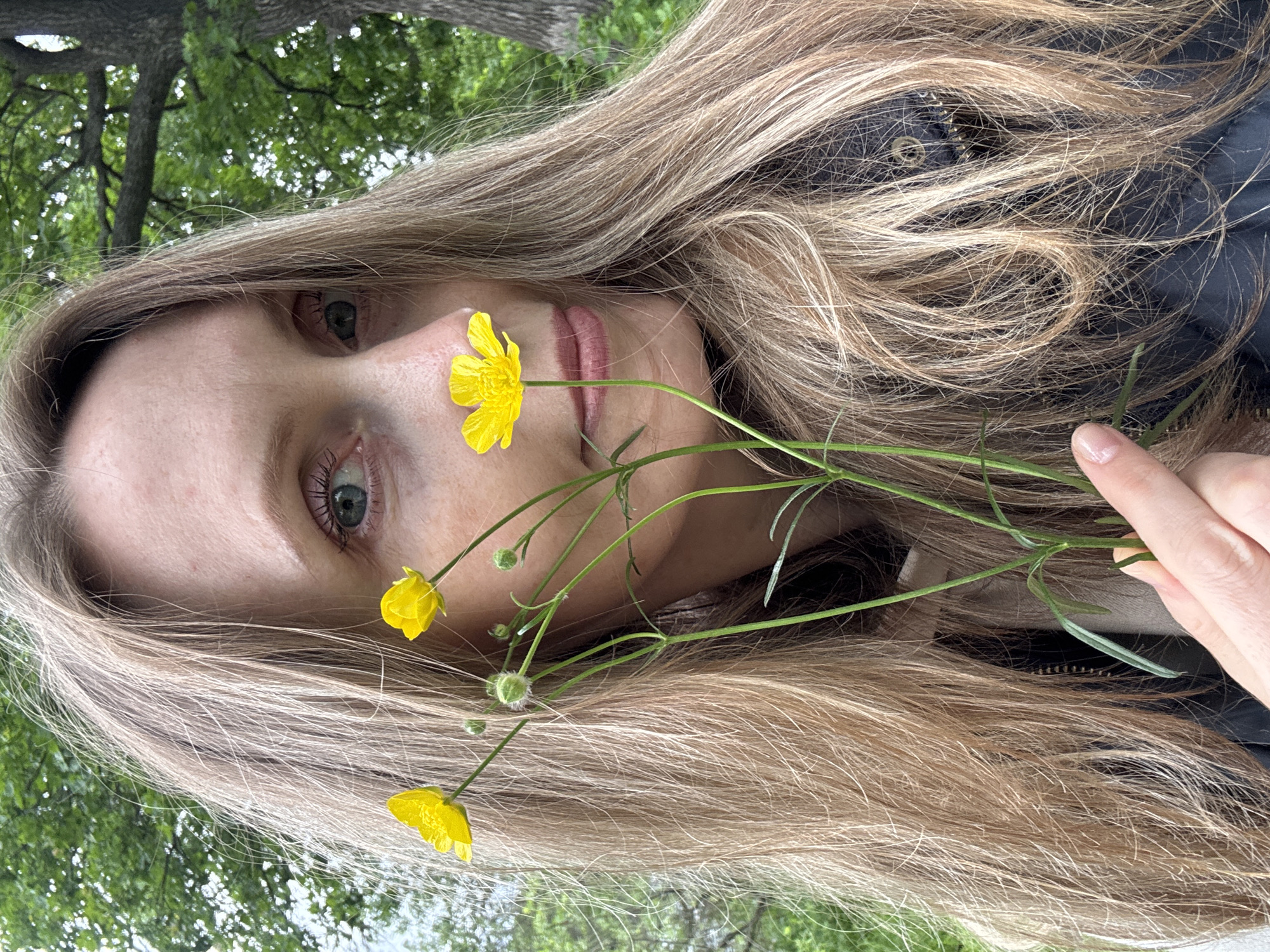 A fair-skinned individual, identified as female, is depicted from the chest up in an outdoor setting in the city of Chaikovsky, Russia. She has long, light brown hair that falls over her shoulders and chest, and light blue-green eyes. Her gaze is directed towards the viewer, and her lips are slightly parted in a gentle expression. She is wearing a dark-colored jacket with a visible zipper, over a lighter, possibly beige, garment. In her right hand, she holds a small cluster of buttercup flowers, characterized by their bright yellow petals and green stems, along with several closed buds. One flower is positioned near her nose, another near her left eye, and a third to the left of her face. The background is blurred, featuring dense green foliage, indicative of trees and bushes, with a tree trunk visible on the right side. The lighting suggests an overcast or shaded natural environment.