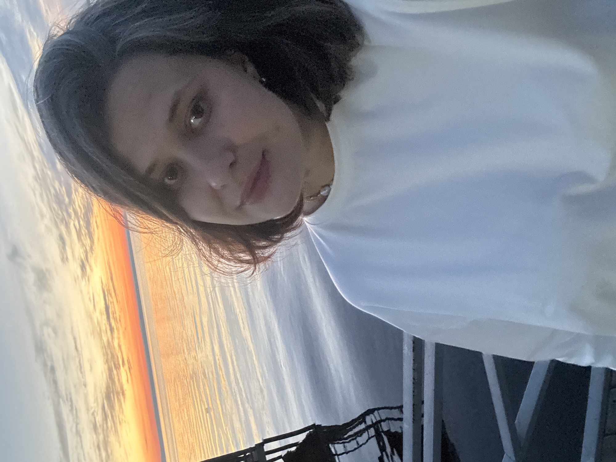 The image is a selfie taken by a young woman outdoors during a sunset. She has dark hair and is wearing a white t-shirt. The background shows a body of water with the sky transitioning from orange and yellow at the horizon to a paler sky above. A dark structure, possibly a pier or railing, is visible on the left side of the frame. The overall lighting suggests it is either sunrise or sunset. The location provided, Костоваты, Russia, is not discernible from the visual elements of the image itself.