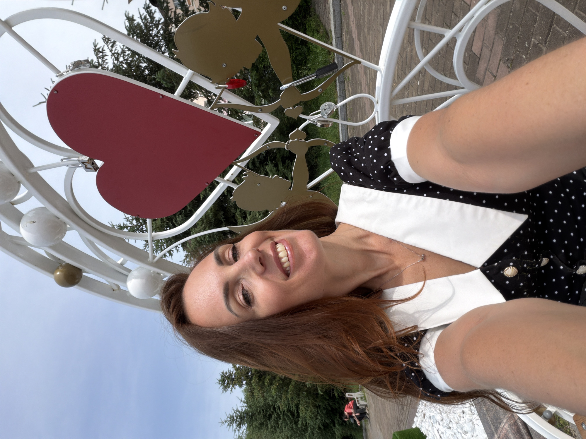 A woman with long, auburn hair is taking a selfie outdoors. She is smiling and wearing a black and white polka dot dress with a white collar and a silver necklace. Behind her is a large white heart-shaped arch with a red heart in the center. Two golden cupid figures with bows and arrows are part of the arch's decoration, and several padlocks are attached to it. In the background, there are evergreen trees and a pathway. Two people are sitting on a bench in the distance. The scene appears to be in a park or public space in Birobidzhan, Russia, likely a location where couples place love locks.