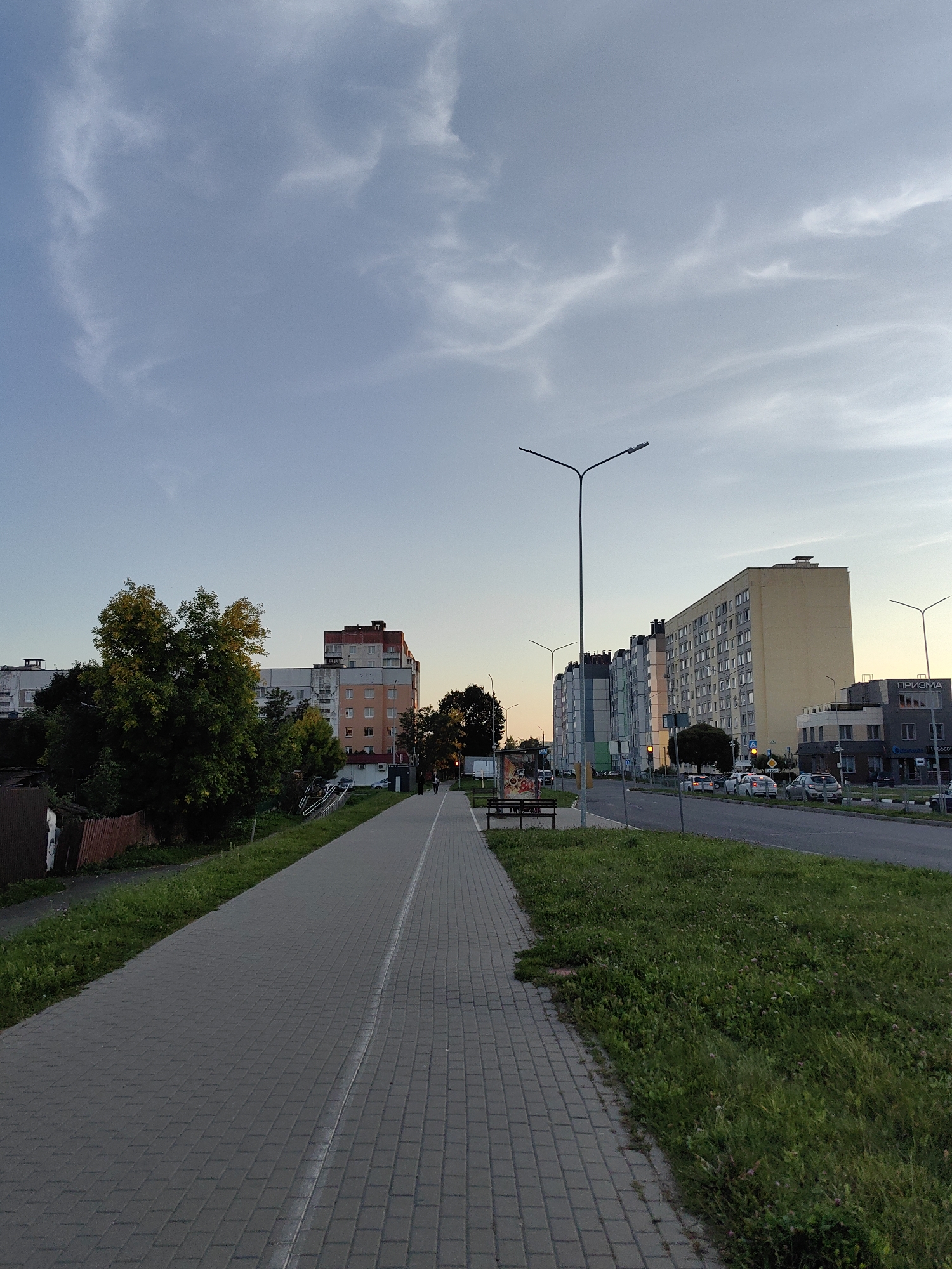 This is a vertical, eye-level, outdoor, daytime shot of a paved walkway running alongside a street in Fanipal, Belarus. The sky is a pale blue with wispy white clouds. The walkway is made of gray pavers and has a white line marking its edge. To the left of the walkway, there is a patch of green grass and some trees with yellowing leaves, suggesting it is autumn. Further left, a corrugated metal fence and the side of a building are visible. To the right of the walkway, the street is lined with several multi-story apartment buildings of various architectural styles and colors. The buildings vary in height and some appear to be newly constructed. Cars are parked along the street and some are in motion. A bus stop shelter with a bench is present on the left side of the walkway. Two individuals are walking on the walkway in the distance, and a few more are visible on the sidewalk across the street. Streetlights are visible on both sides of the street. The overall impression is one of an urban residential area.