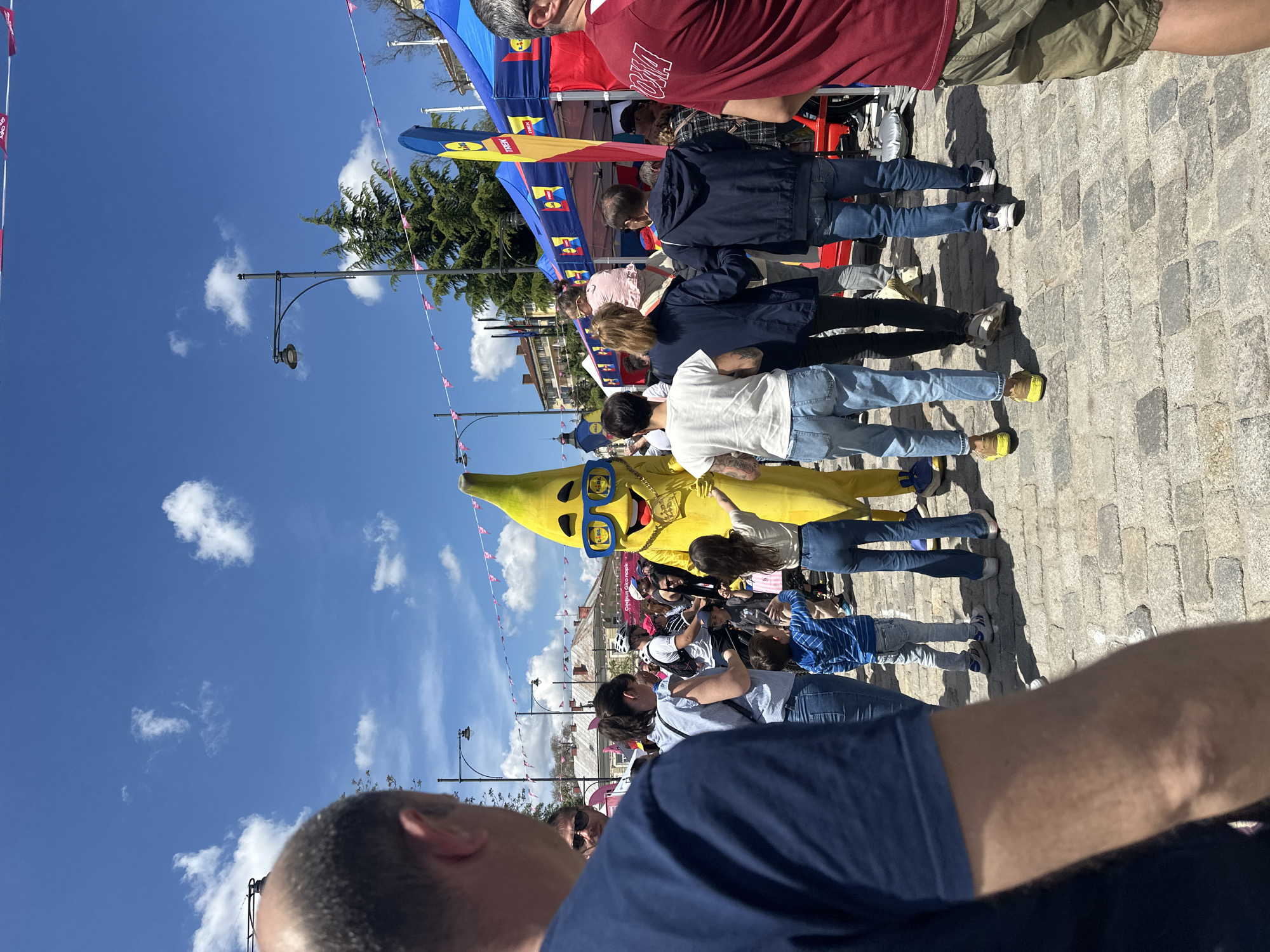 The image shows a crowd of people gathered on a cobblestone street in what appears to be a public event or festival. The sky is blue with scattered clouds, and buildings are visible in the background. A person dressed in a banana costume with blue sunglasses and a gold chain is interacting with the crowd, primarily children. Overhead, there are banners and flags strung between streetlights. People are standing and looking towards the banana character, some taking photos. The image seems to be taken from a low angle, with part of a person's head and shoulder visible in the foreground. Although the location cannot be confirmed to be specifically in Sofia, Bulgaria without further evidence from the image, the presence of European architecture is apparent.