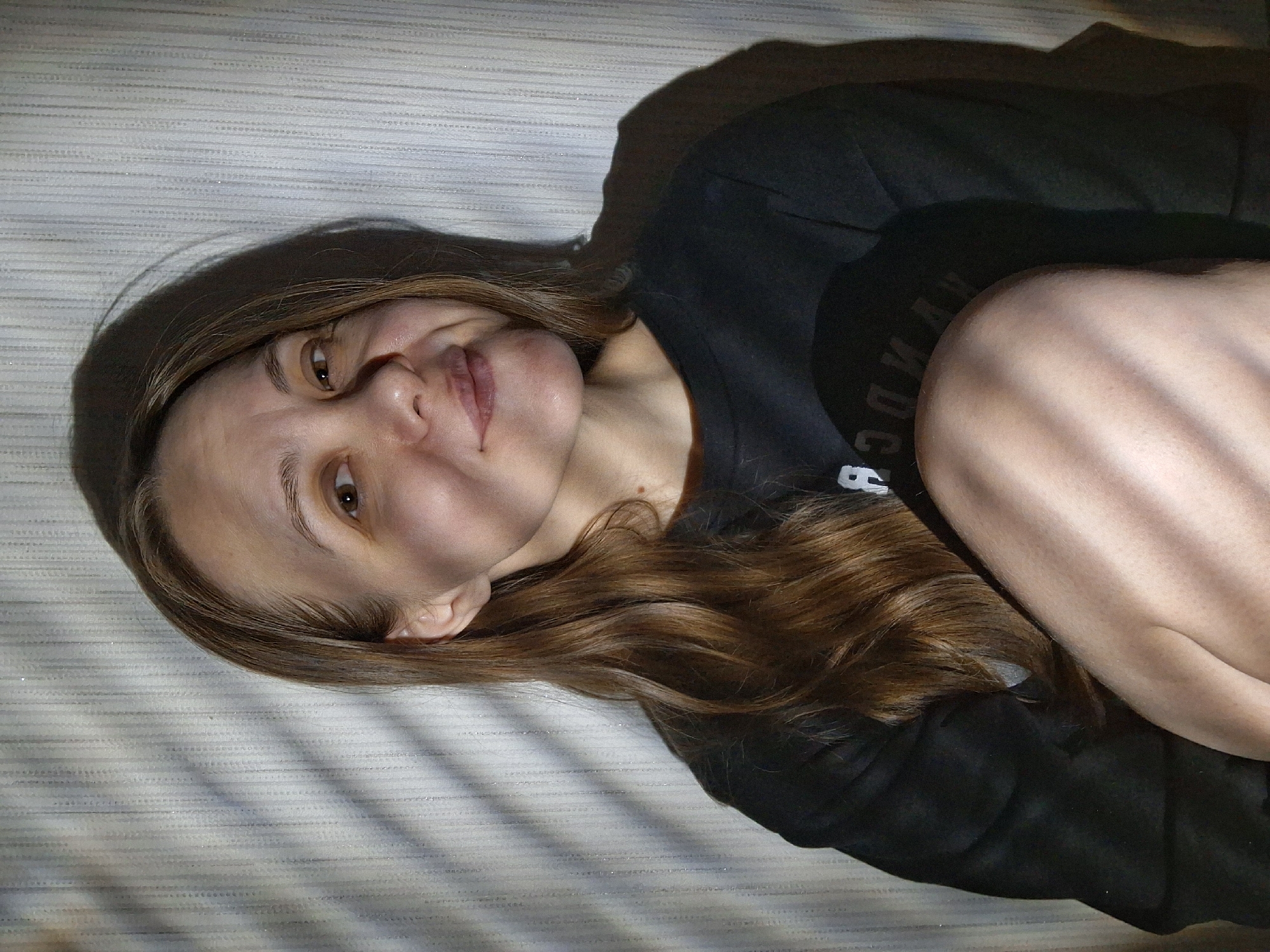 A woman lies on a horizontally striped surface, likely a bed or similar furniture. She wears a black long-sleeved shirt. Her hair is long, wavy, and brown, with some lighter highlights. She is facing the camera with a slight smile. Her skin appears pale, and light patterns cast shadows across her face and the striped surface. Part of her arm or leg is visible near her shoulder. The image suggests an indoor setting. The location of Chaikovsky, Russia, is not visually discernible from this image.