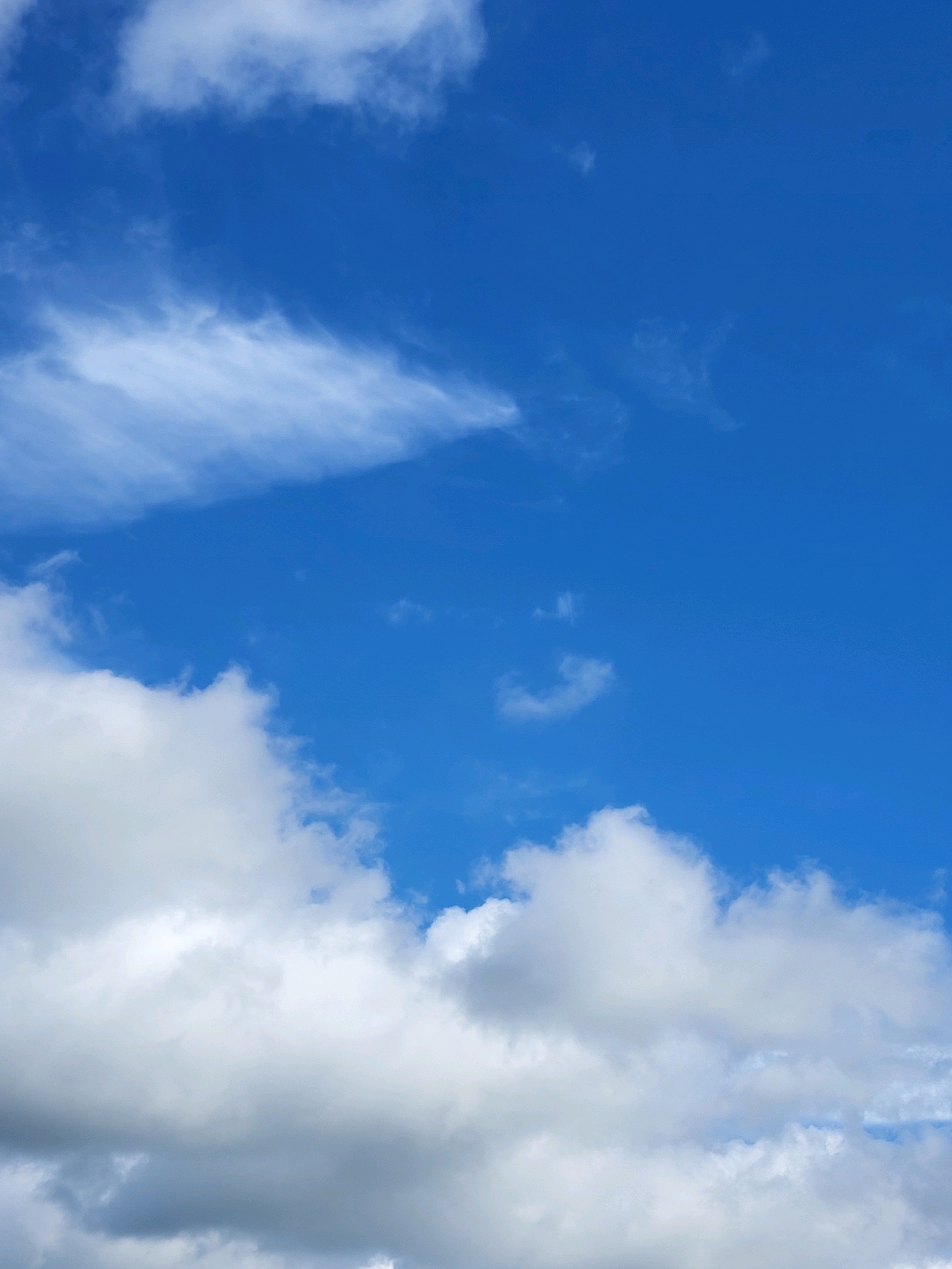 The image shows a bright blue sky with scattered clouds. In the foreground, at the bottom of the frame, are large, puffy white and grey clouds. Higher up, there are wispy, thinner clouds stretched across the sky. The overall impression is of a clear, partly cloudy day. There are no people, objects, or discernible landmarks visible, nor any indication of a specific location within a city.