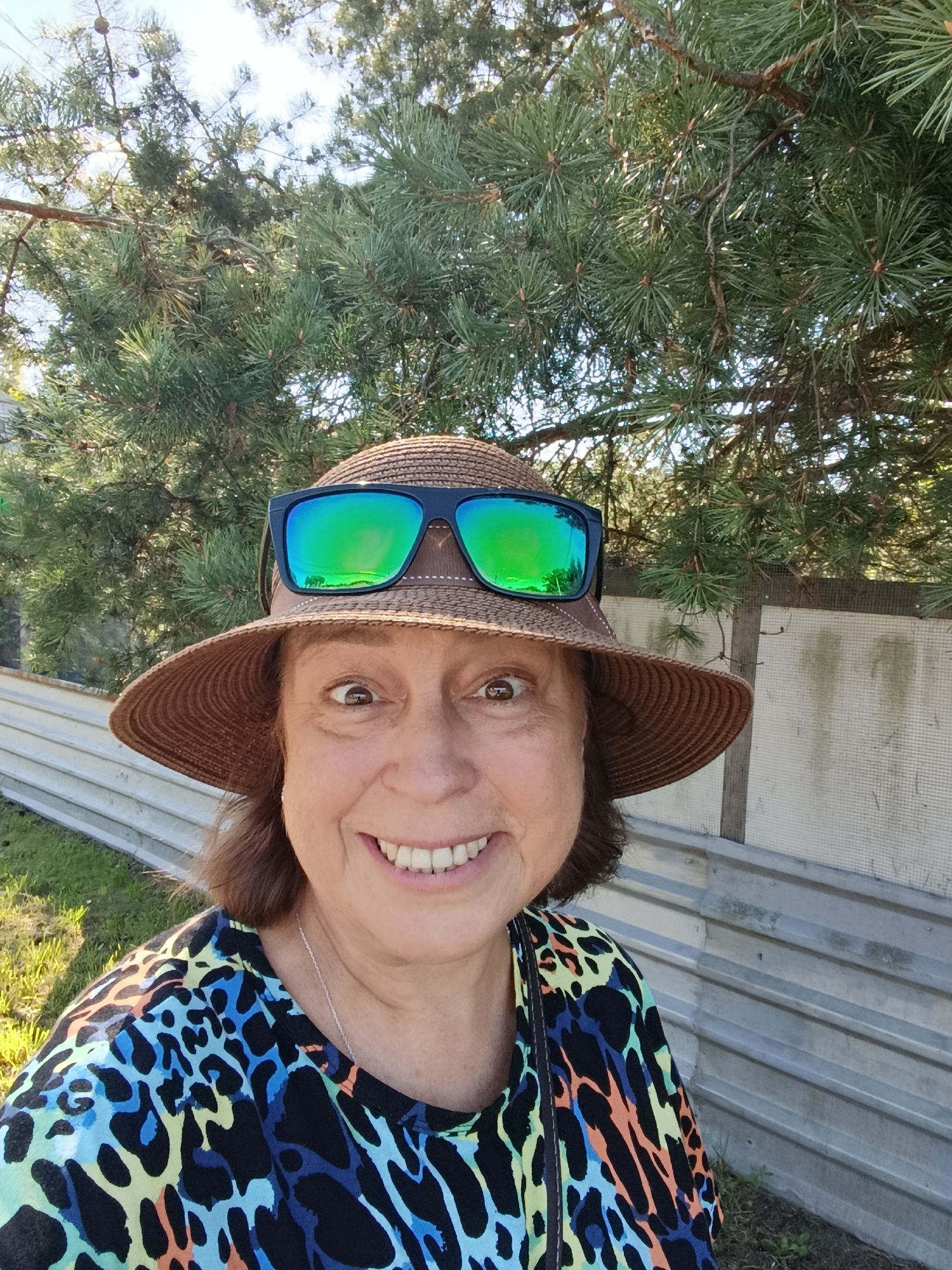 A woman wearing a straw hat and sunglasses is taking a selfie outdoors. She has brown hair and is smiling. She is wearing a colorful top with a leopard-like print. Behind her, there are pine trees and a corrugated metal fence. The scene appears to be in a suburban or rural area. The image is taken during the daytime, with sunlight visible. No specific location in Vareya, Russia, can be identified from the image.