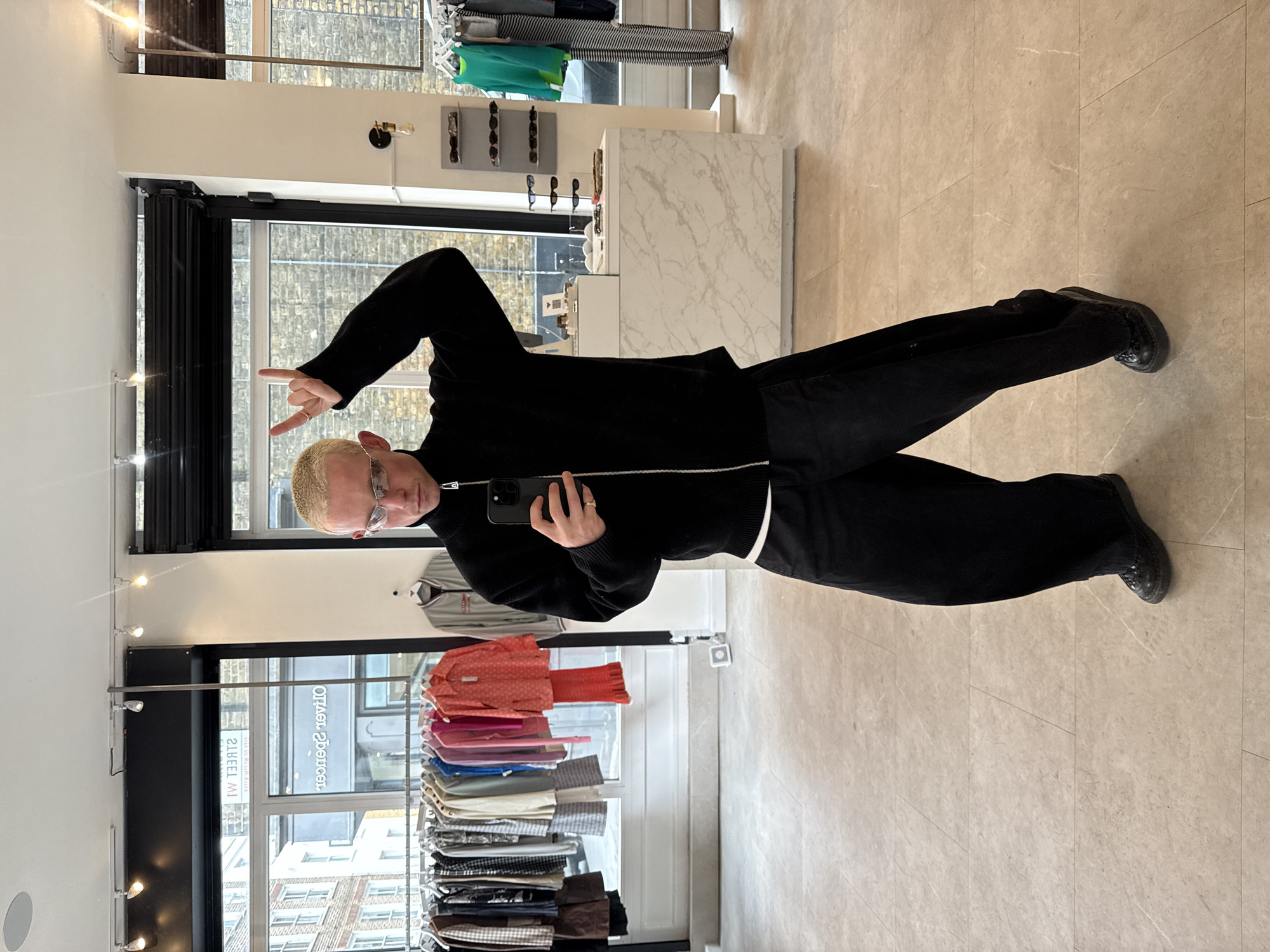 A man is taking a selfie in a shop in London, United Kingdom. He is wearing a black jacket, black pants, and black shoes. He is holding a black smartphone in his right hand and making a peace sign with his left hand. The shop has clothing racks with various garments. The shop windows show the city landscape outside. The floor is tiled with a light beige pattern.