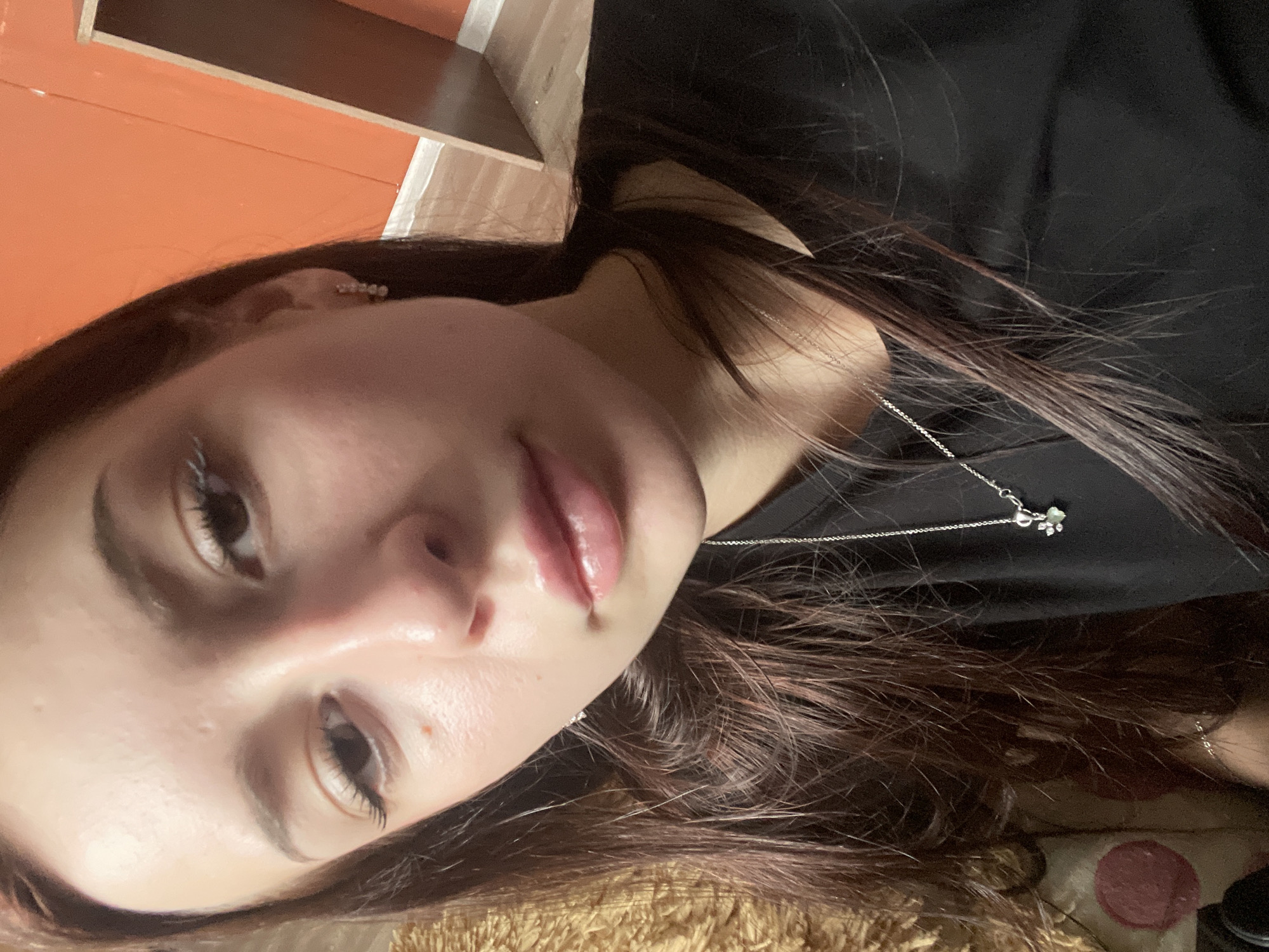 A young woman with long, dark brown hair is captured in a close-up selfie. She is looking towards the camera with her head slightly tilted. Her eyes are brown and appear to have white mascara on her lashes. She is wearing a black t-shirt and a silver necklace with a small, detailed pendant. The background shows an orange wall and a wooden structure, suggesting an indoor setting. The lighting is soft, casting a gentle glow on her face. There is a hint of a textured, yellowish surface to the left of the frame, possibly a piece of furniture or fabric. The image does not contain any information to suggest a specific location in the city of Медведево, Russia.