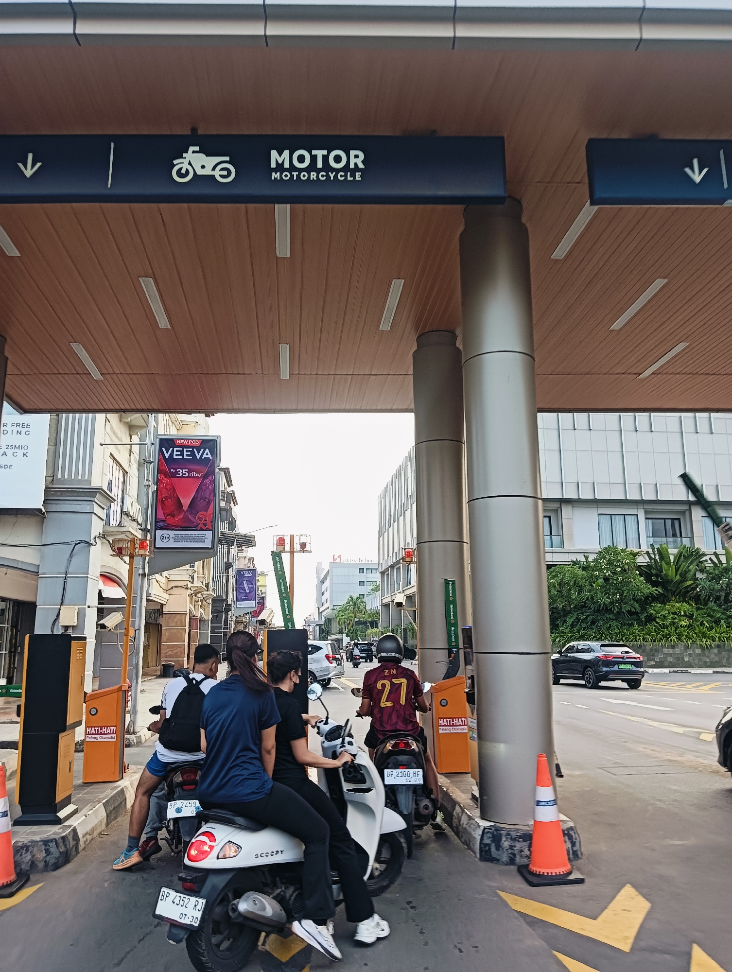The image shows a street scene at what appears to be an entrance to a paid parking area. In the foreground, three people are seated on two motorcycles. One motorcycle, a white scooter, is in the center, with two individuals riding it. The rider in the back wears a dark blue shirt and black pants, while the rider in the front wears a white shirt and blue shorts and has a black backpack. The second motorcycle, a darker color, is to the left of the scooter, with one person wearing a maroon and yellow jersey with the number 27. The individuals are facing forward, as if waiting to proceed.
Above the motorcycles, a dark blue sign indicates "MOTOR MOTORCYCLE" with a downward-pointing arrow on the left and a similar arrow on the right. The sign is mounted on a wooden-paneled ceiling structure supported by large cylindrical metallic pillars. To the left, yellow parking ticket machines are visible, with "HATI-HATI" (Caution) and "Pulang Otomatis" (Automatic Return) text on them.
In the background, a multi-story building with modern architecture is visible on the right, featuring a facade of glass and white panels. Green trees and foliage are present in front of this building. On the left side of the image, other buildings with storefronts and advertising billboards are present. One prominent billboard displays "VEEVA" with a price of "Rp 35 ribu". Traffic cones are placed on the ground near the ticket machines. The overall lighting suggests it is daytime. The road surface has yellow lane markings.