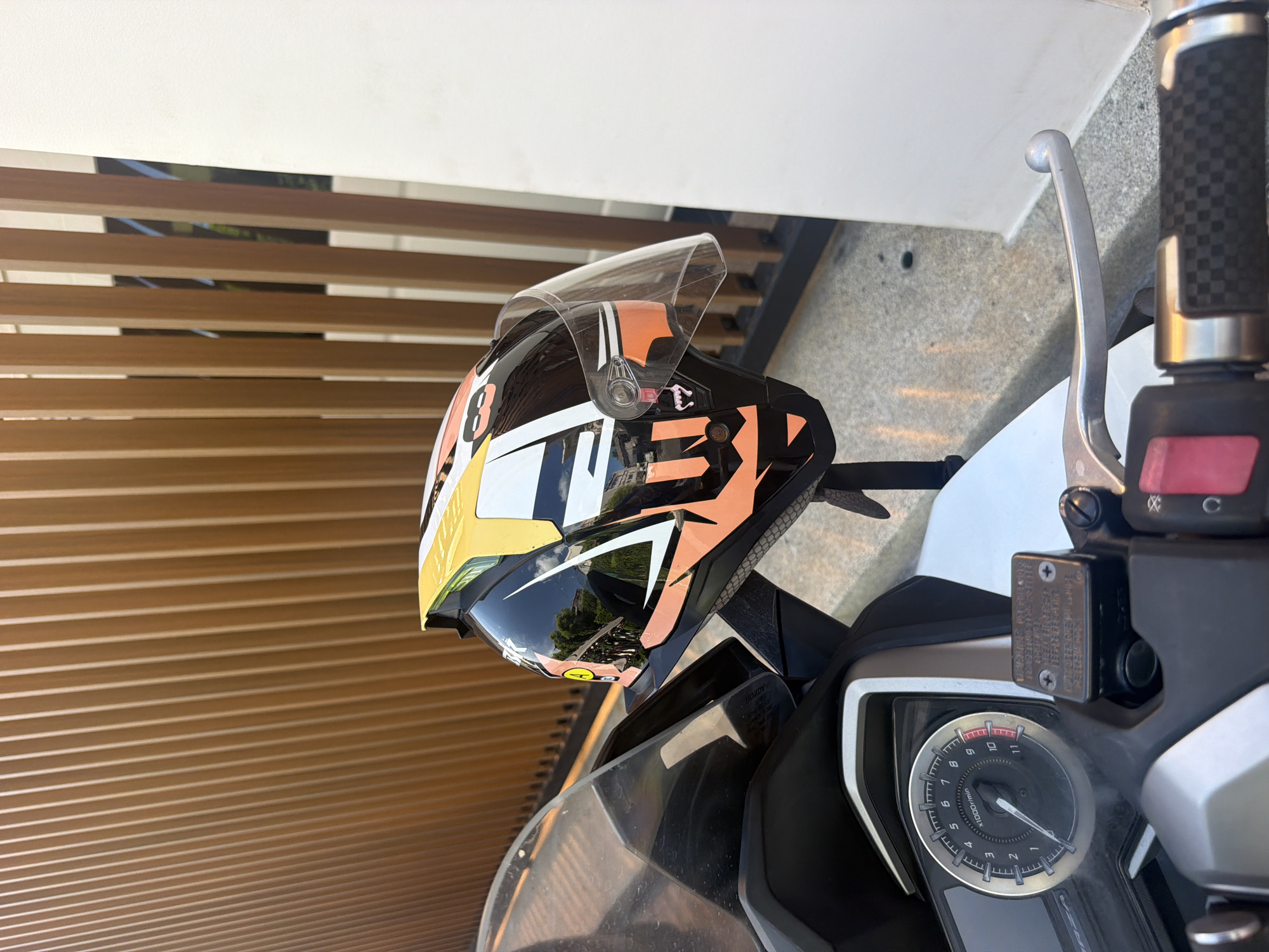 A close-up, eye-level shot shows a black motorcycle helmet with a clear visor, decorated with orange and white graphics, resting on the front of a scooter. The helmet is positioned to the right of the scooter's windscreen, which is visible on the left side of the frame. A portion of the scooter's dashboard, including a circular tachometer with a needle pointing between 9 and 10, is visible in the lower left corner. To the right of the helmet, a wooden slatted fence is visible, with a white wall to the far right. The ground appears to be a paved surface with small stones. The lighting suggests it is daytime, casting shadows. The scene is located in Baan Chaweng, Thailand.