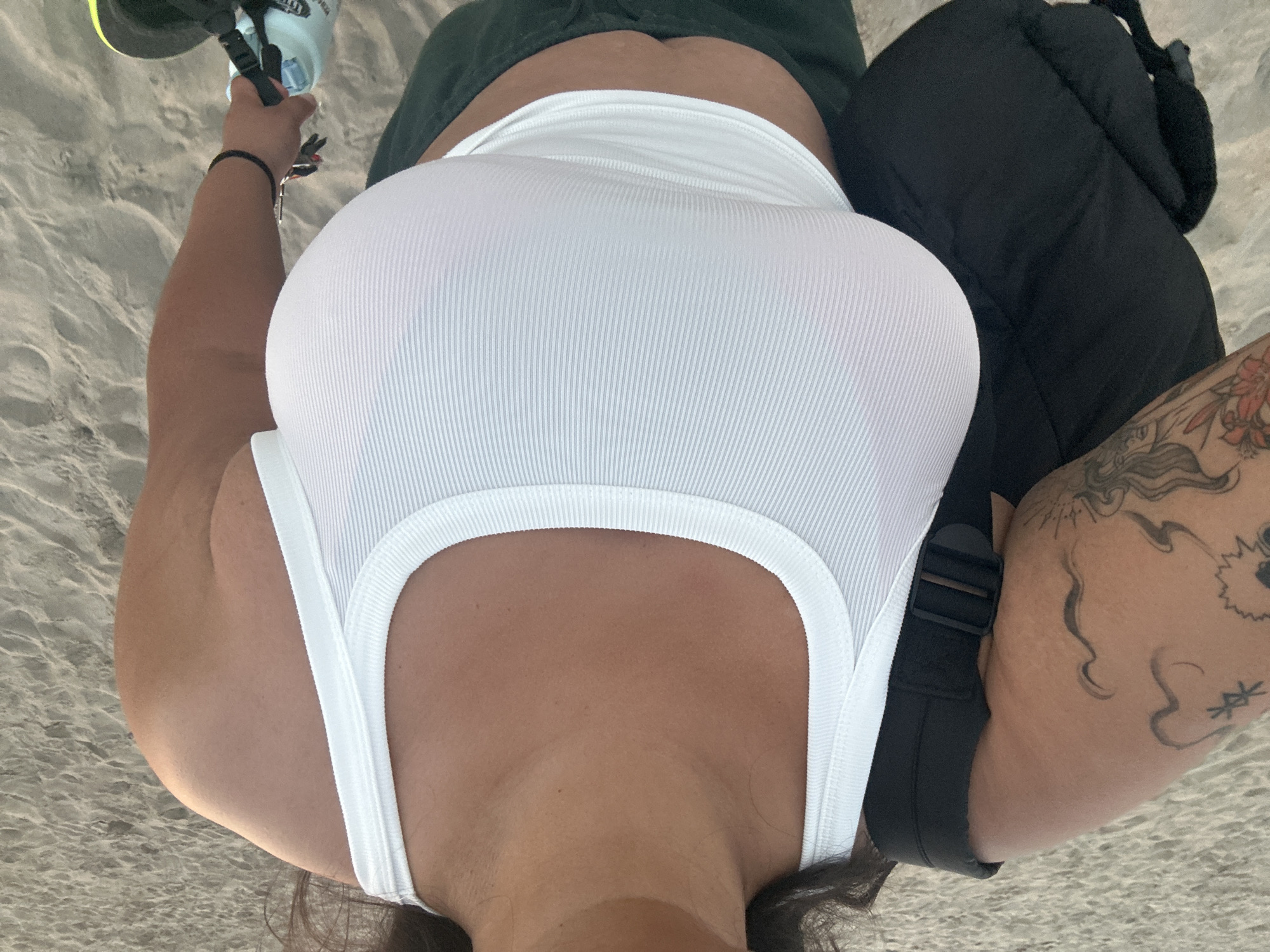 The image is a close-up, top-down shot of a person from the chest up, with the camera angled downwards. The person is wearing a white, ribbed tank top. To the left of the person, their arm is visible, holding a water bottle with a black strap and some keys. Their skin has a tan line across the chest and neck. To the right, a black backpack strap is visible, and part of a tattooed arm with swirling designs is also visible. The background is a sandy beach, indicating an outdoor setting. The lighting suggests it is daytime. The overall impression is one of casual relaxation on a beach.