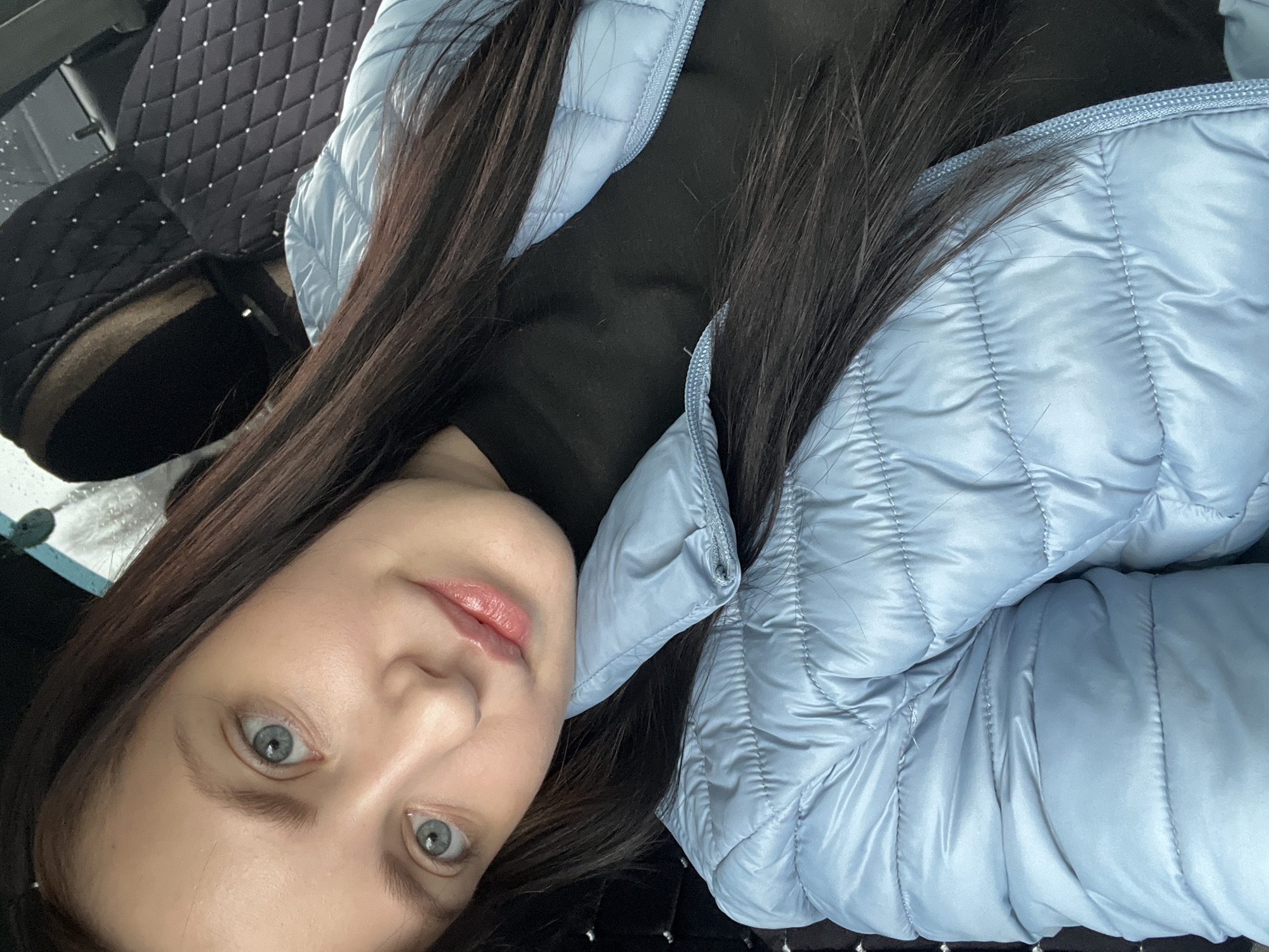 A woman with long dark hair and light blue eyes is lying down, wearing a light blue puffer jacket over a black top. She is indoors, possibly in a car, as a black quilted surface with small white dots is visible in the background. The location is Марковский, Russia.