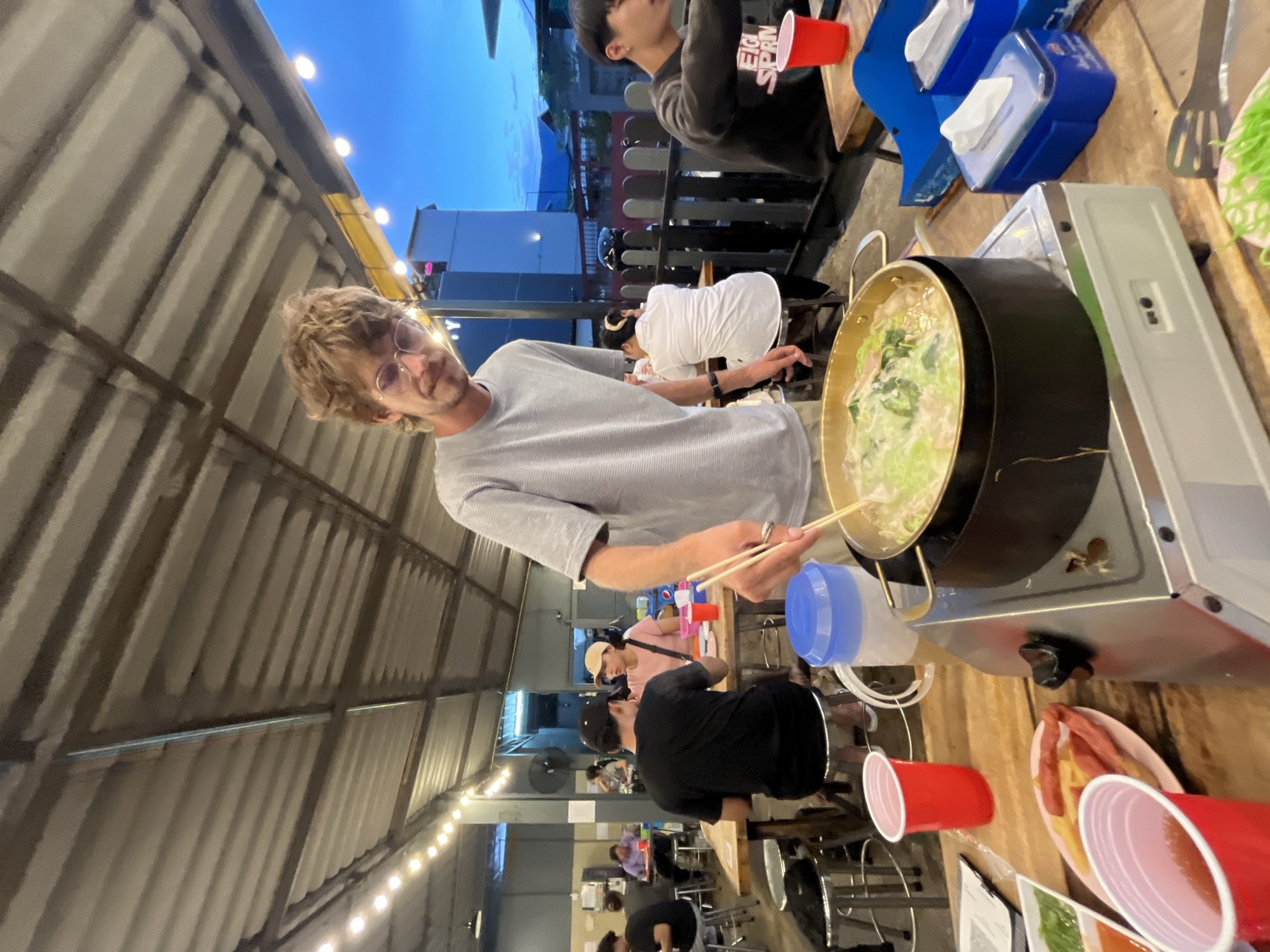 A man with blonde hair and glasses is cooking at a table in what appears to be an outdoor food stall or restaurant in Chiang Mai City Municipality, Thailand. He is wearing a grey t-shirt and is holding chopsticks, stirring a pot of soup filled with vegetables. The pot is on a portable stove. In the foreground, there are red cups, plates of food including what looks like fried items and green vegetables, and a large blue water jug. In the background, other people are seated at tables, eating and socializing. The setting is casual, with wooden tables and chairs, a covered roof overhead with string lights, and a fence in the distance. The sky is visible and appears to be dusk.