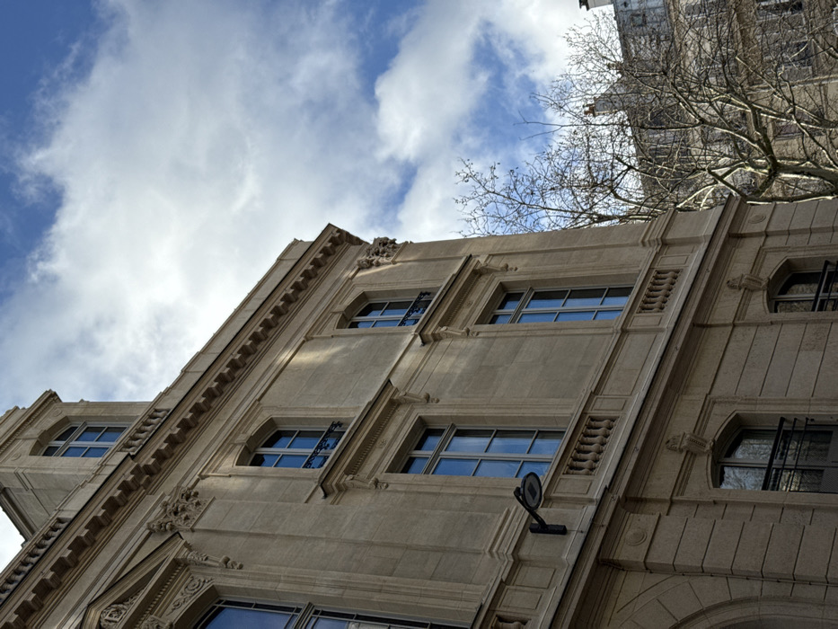 The image displays the facade of a classical Haussmannian-style building, typical of Paris, France, viewed from a low, upward-angled perspective. The structure, constructed from light beige cut stone, features multiple stories with tall, rectangular windows. The window panes reflect a bright blue sky partially obscured by scattered white cumulus clouds. Architectural details include cornices, decorative pilasters, and carved ornamentation around windows and ledges. Several windows incorporate dark wrought-iron Juliet balconies or guards. A black, contemporary street lamp with a flat, circular head is mounted on the building's facade. The uppermost visible section of the building includes an attic level with a prominent, arched dormer window. To the right, bare, dark branches of deciduous trees are visible against the sky and a second, more distant Parisian building with a grey roof and multiple chimneys. No individuals or specific actions are depicted.