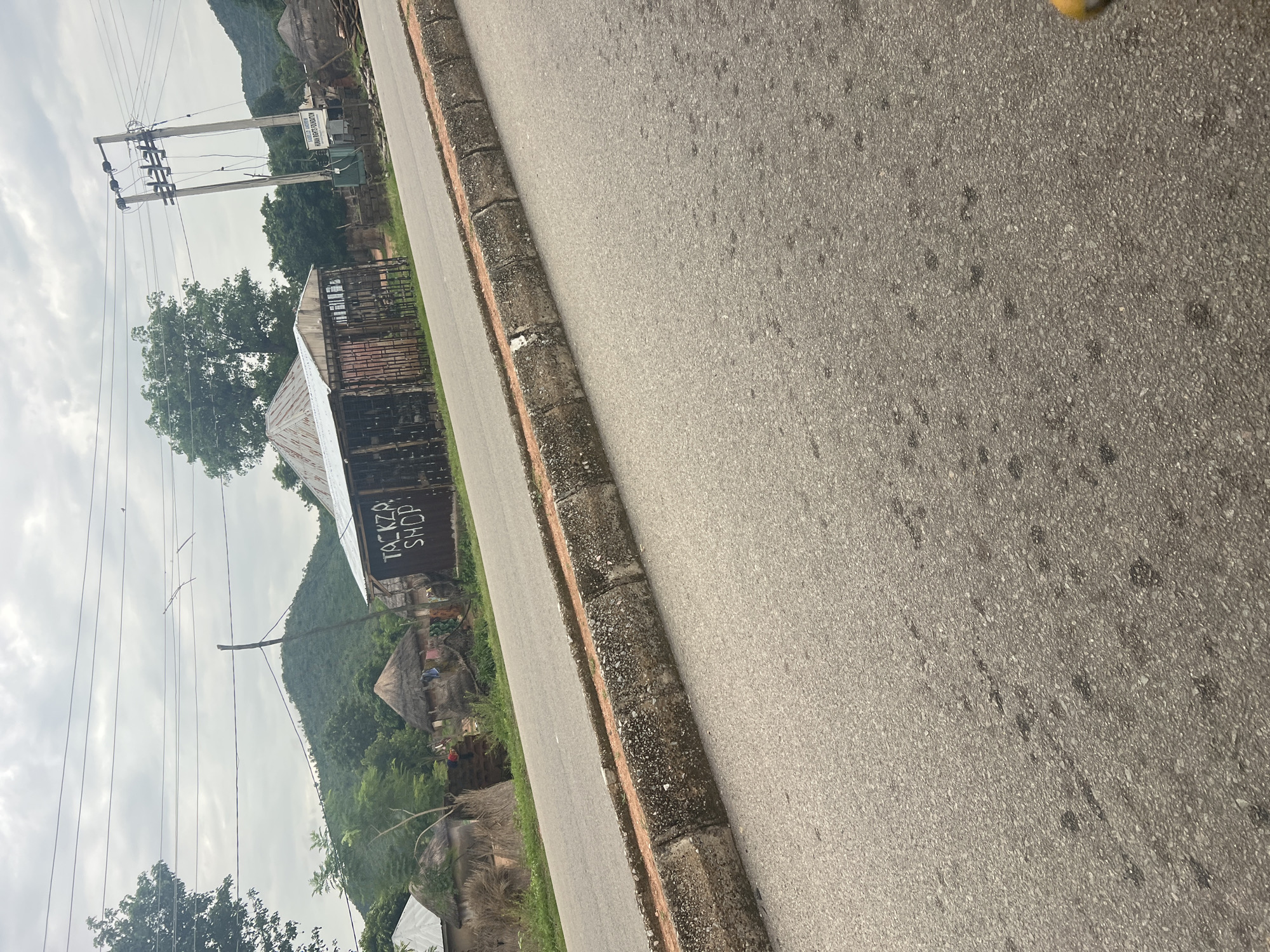 The image shows a rural scene in Kona Garu, Nigeria. A paved road runs diagonally through the frame, with a concrete and stone curb separating it from a grassy area. On the left side of the road, there are several buildings. One building has a corrugated metal roof and appears to be a shop, with the text "TOKIZA SHOP" painted on its side. Next to it are traditional thatched-roof huts. Beyond the buildings, a lush green hillside rises, dotted with trees. Overhead, power lines stretch across a cloudy sky. The overall impression is of a quiet, everyday scene in a developing community. The lighting suggests it is daytime, possibly overcast.