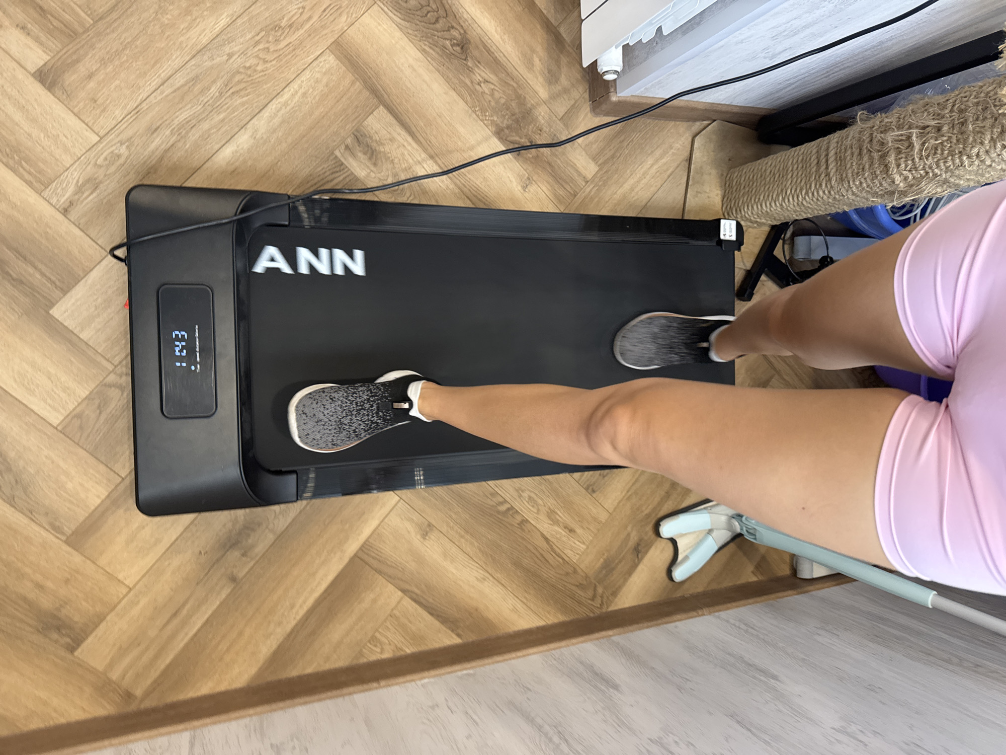 A person's legs are visible from the knees down, wearing pink shorts and gray and black athletic shoes. They are standing on a black treadmill with a digital display showing "12:43". The treadmill is on a wooden floor with a herringbone pattern. To the left of the treadmill, a mop with a light blue head and handle is leaning against a wall. To the right of the treadmill, a cylindrical object wrapped in rope is visible, along with a white radiator and a dark blue object. The scene appears to be indoors.