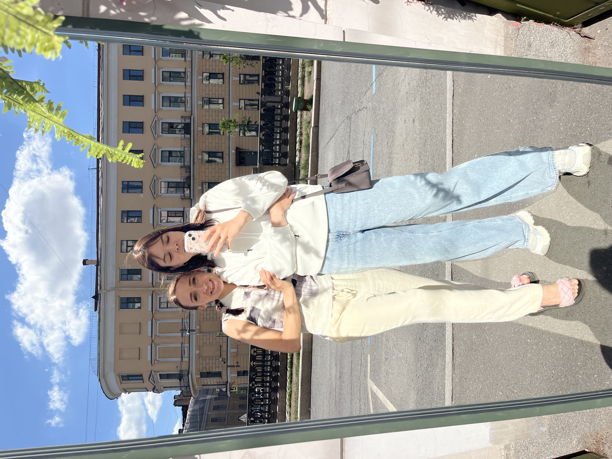 Two young women pose for a mirror selfie on a sunny day in Saint Petersburg, Russia. The woman on the left, with her hair in a braid, smiles at the camera wearing a sleeveless patterned top and light-colored pants with pink slides. The woman on the right, holding a phone to take the photo, wears a white hooded sweatshirt and light blue wide-leg jeans with white sneakers. A dark gray crossbody bag hangs from her shoulder. They are positioned in front of a large mirror that reflects their image and the surroundings. Behind them, a pale yellow building with numerous windows and balconies stands prominently, a typical architectural style found in St. Petersburg. A black wrought-iron fence runs along a sidewalk in front of the building, and a street with cars and pedestrian crossing signs is visible. Greenery, including a lush fern frond partially obscuring the top right of the mirror, adds a touch of nature to the urban scene. The sky is a vibrant blue with scattered white clouds.