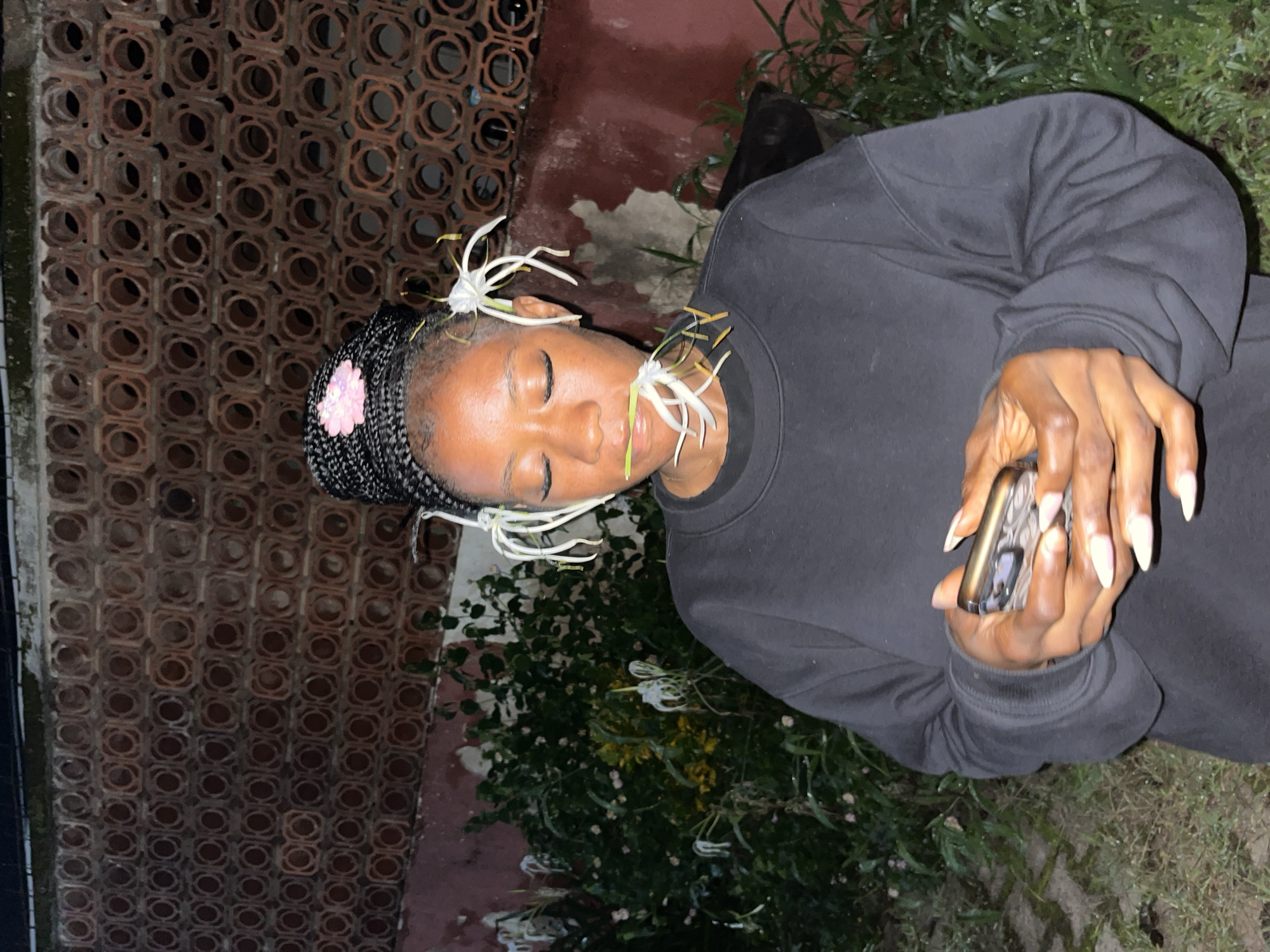 A young Black woman with her eyes closed and flowers in her hair and mouth is holding a phone in a garden at night. She is wearing a black sweatshirt. Behind her is a reddish-brown brick wall with a circular pattern and lush green vegetation. She has long, light-colored fingernails. The setting appears to be an outdoor area, possibly a backyard or garden.