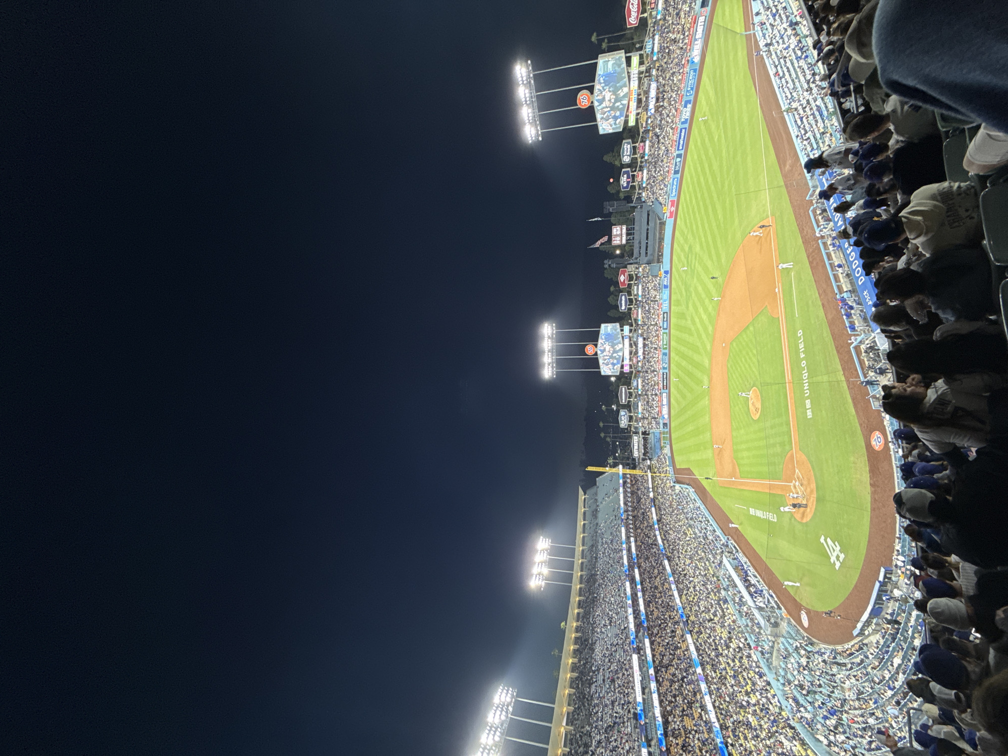 The image shows a baseball game at Dodger Stadium in Los Angeles, California, at night. The stadium is filled with spectators, most of whom are wearing blue, the color of the Los Angeles Dodgers team. The baseball field is green and brown, and is well-lit by stadium lights. Advertisements are visible around the stadium, including for Coca-Cola, UNIQLO FIELD, and drum. There are large screen displays, one of which shows "76" on it. Several people are seated nearby, their heads and shoulders visible in the lower right of the frame. The night sky is visible in the upper left portion of the image.