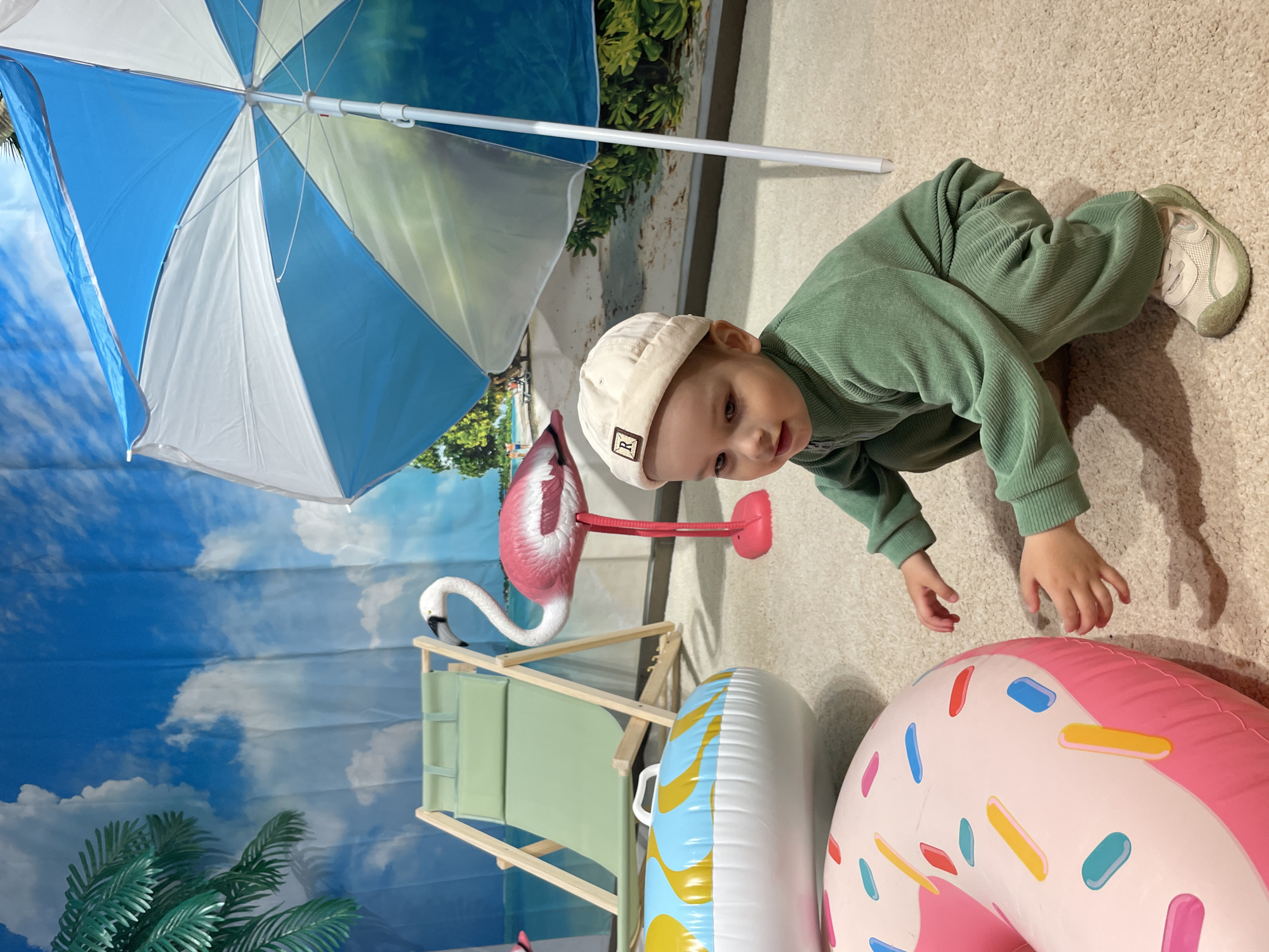 The image shows a young child in a green outfit and a light-colored hat. The child is on a beige carpet and appears to be reaching towards a pink inflatable donut. Next to the donut is a white and blue inflatable tube. Behind the child are a small green beach chair and a plastic flamingo. Above these objects is a blue and white beach umbrella. The background is a beach-themed backdrop featuring a blue sky, clouds, and palm trees. The location is Unknown, Unknown.