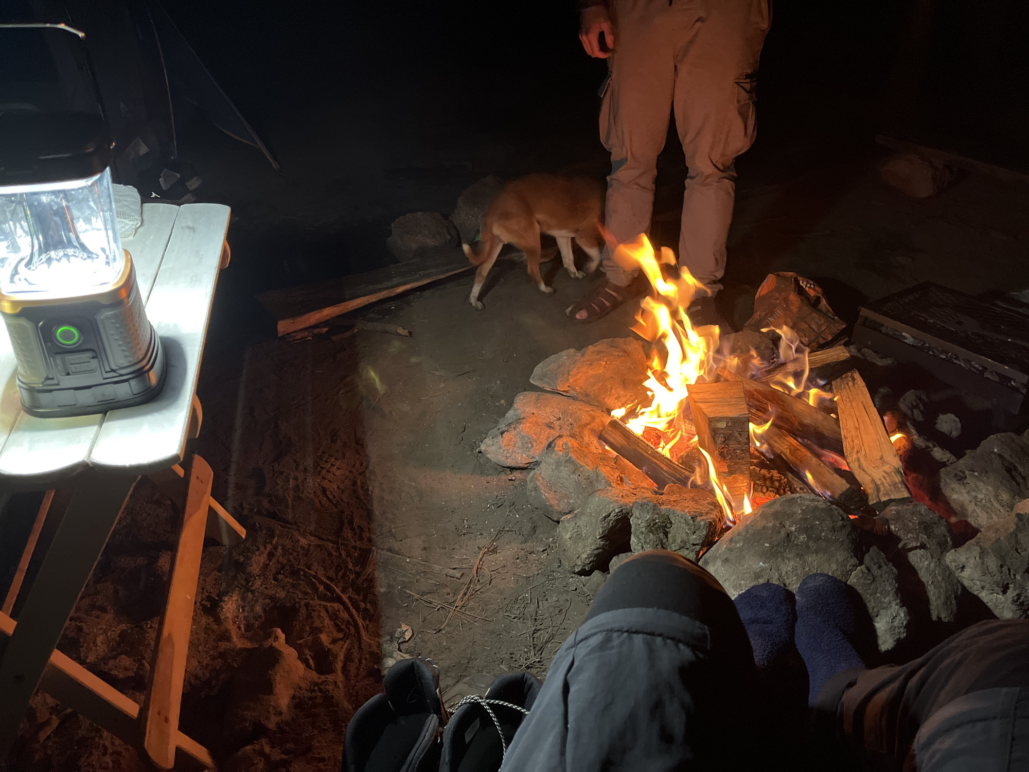 The image depicts a nighttime scene at a campsite in El Paisano, Mexico. A campfire crackles in the center, surrounded by rocks and dirt. To the left, a white and wooden picnic table holds a portable lantern. In the background, a person in tan cargo pants stands near the fire, while a small brown dog sniffs the ground. In the foreground, two pairs of feet, one in black shoes and the other in blue socks, are visible, suggesting people are sitting around the campfire. The environment is dark, illuminated primarily by the campfire and lantern.