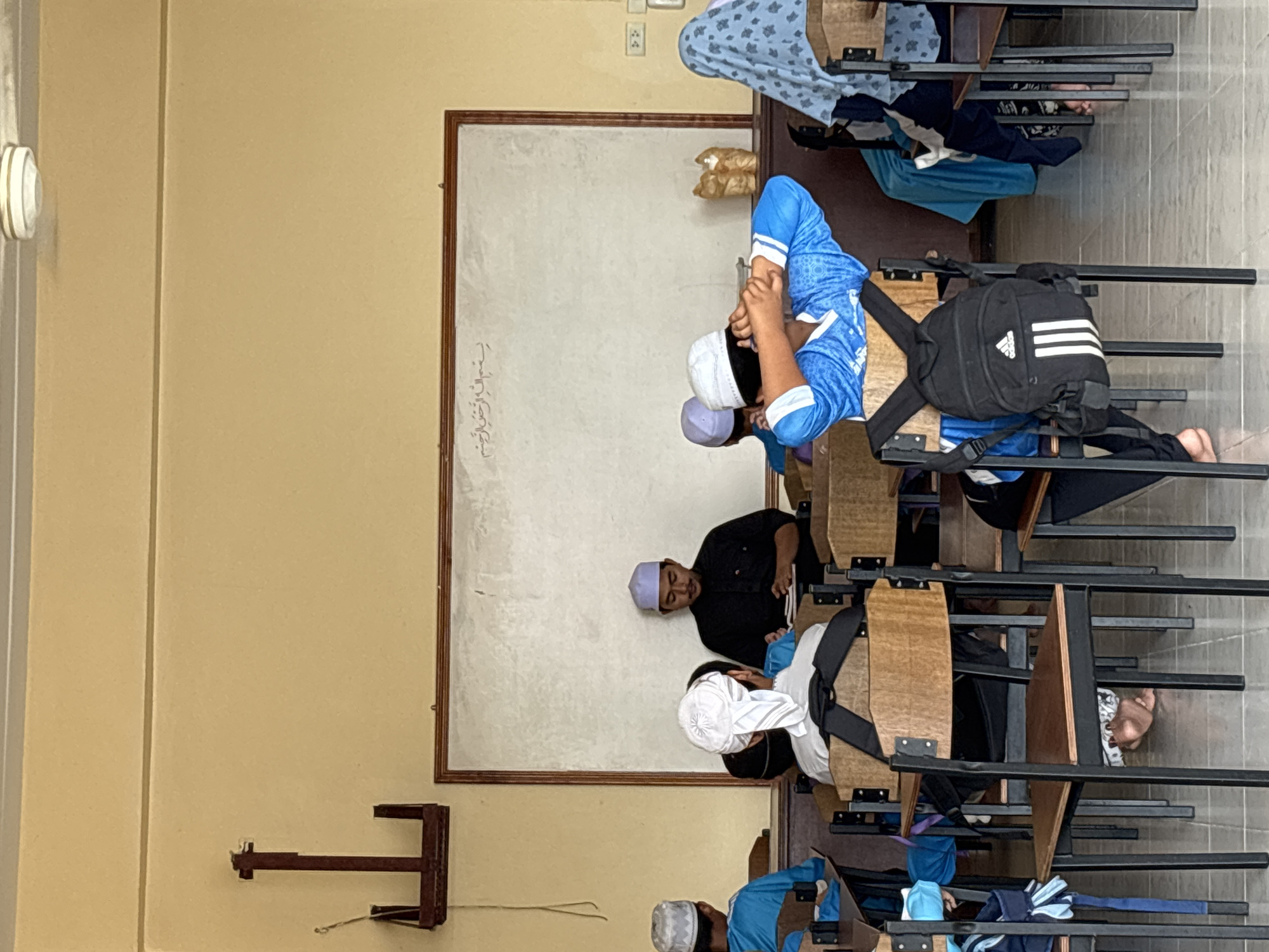 The image depicts a classroom scene, likely in Rawai, Thailand, where a group of young males, dressed in traditional Muslim attire including kufis and tunics, are seated at desks. A male figure, presumably a teacher, is at the front of the classroom, facing a whiteboard. The whiteboard has Arabic inscriptions at the top. The classroom has pale yellow walls and a tiled floor. There are several wooden chairs with metal frames, some with backpacks attached. Some students are looking forward, while others have their heads down or are turned away. One student in a blue shirt is stretching his arms behind his head. Another student's feet are visible under the desk. A decorative wooden object is placed on a shelf behind the students. The lighting suggests it is daytime.