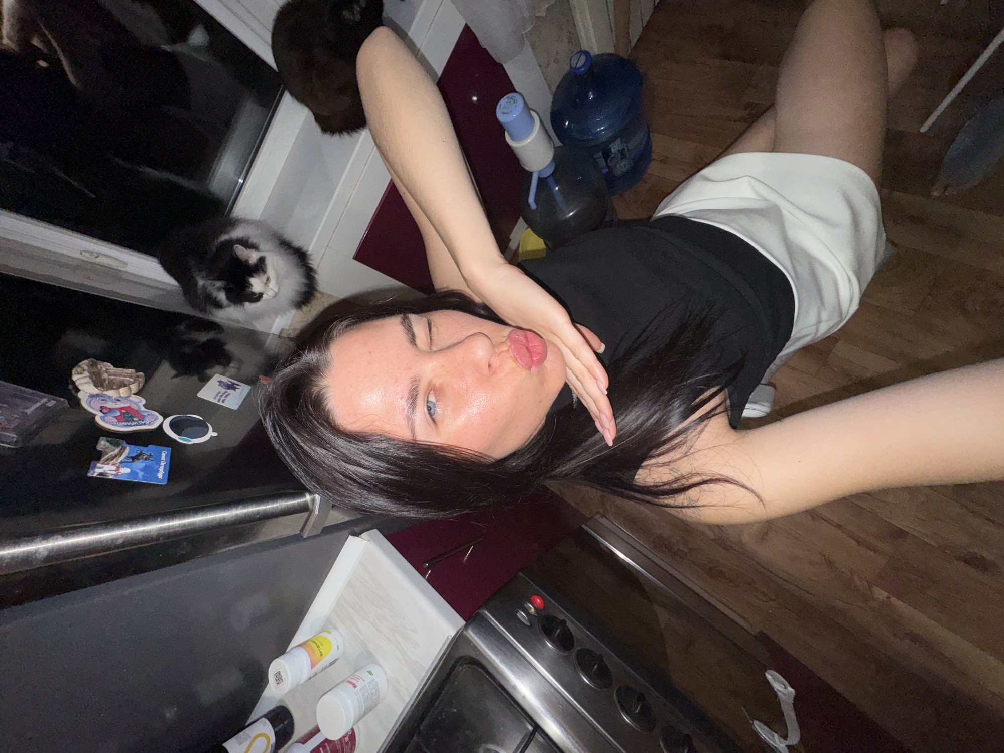 A young woman with long dark hair and blue eyes winks at the camera while making a kissing gesture. She is wearing a black sleeveless top and a white skirt. She is in a kitchen, with a stove visible to her left. On a shelf above the stove are several bottles and jars. A refrigerator is to her left, adorned with various magnets and stickers. In the background, near a window, are two cats, one black and white, and another dark-colored cat. Several large water bottles with dispensers are visible in the lower right portion of the image. The floor is made of wooden planks.