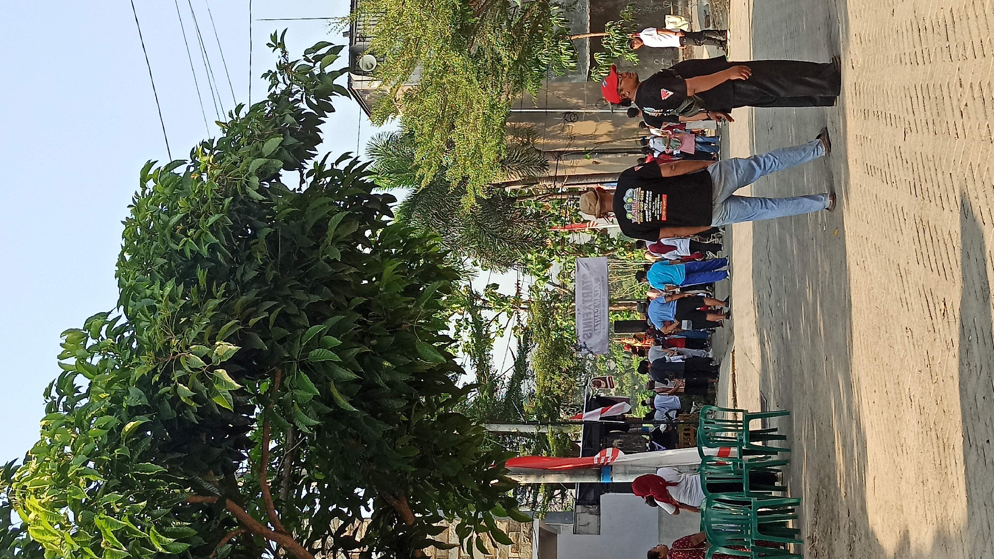 In the city of Galak, Indonesia, a group of people are gathered on a dusty street. The street is lined with green plastic chairs on the left and trees with lush green foliage on the right, casting dappled shadows. In the background, a banner hangs across the street, displaying text in reverse. A few individuals stand out: one man in a black t-shirt with a graphic print and light blue jeans, facing away from the camera, and another man in a black t-shirt and black pants, wearing a red cap, standing to his right. Several other people are milling about, some in casual attire, while a few appear to be organizers near the banner. A red and white Indonesian flag is visible on a pole to the left. The sky is clear and bright, suggesting it is daytime. Buildings with light-colored walls are visible in the background, interspersed with more trees. The scene appears to be a public gathering or event.