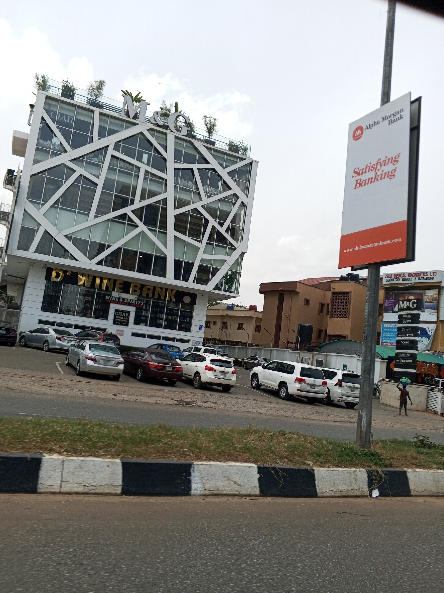 The image depicts a street scene in Abuja, Nigeria. The prominent building is a multi-story structure with a distinctive white frame facade and glass windows. The signs on the building indicate it houses "D' Wine Bank" (wine and spirits) and a restaurant called "M&G." A parking area in front of the building contains several parked vehicles, including sedans and SUVs.
To the right of the building is a tall sign for "Alpha Morgan Bank" and the sign for "Zeda Medical Diagnostics Ltd", another commercial sign advertises for "KINABS", a "scientific equipment consumerables and services" company and "perfumes". A man carrying an object on his head walks along the sidewalk. A road with a black and white curb separates the viewer's perspective from the immediate area. The sky is overcast.