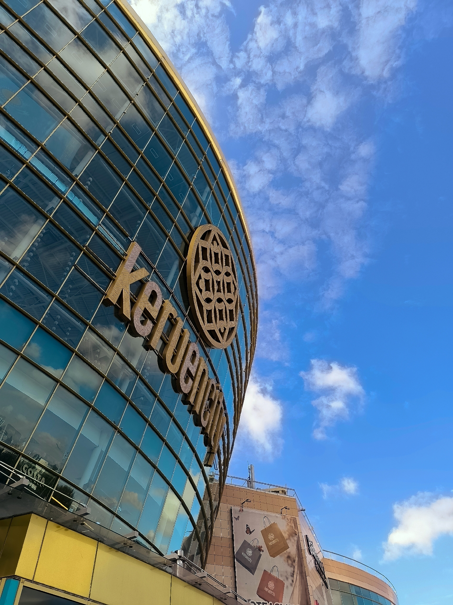 The image shows the Khan Shatyr Entertainment Center in Astana, Kazakhstan, viewed from a low angle, looking up at its distinctive curved glass facade against a bright blue sky with scattered white clouds. The golden lettering "KERUEN" is prominently displayed on the building. Below the curved section, a bright yellow lower section of the building is visible. To the right, a smaller building with banners depicting shopping bags and butterflies is attached. The reflection of the sky and clouds is visible in the glass panels of the Khan Shatyr.