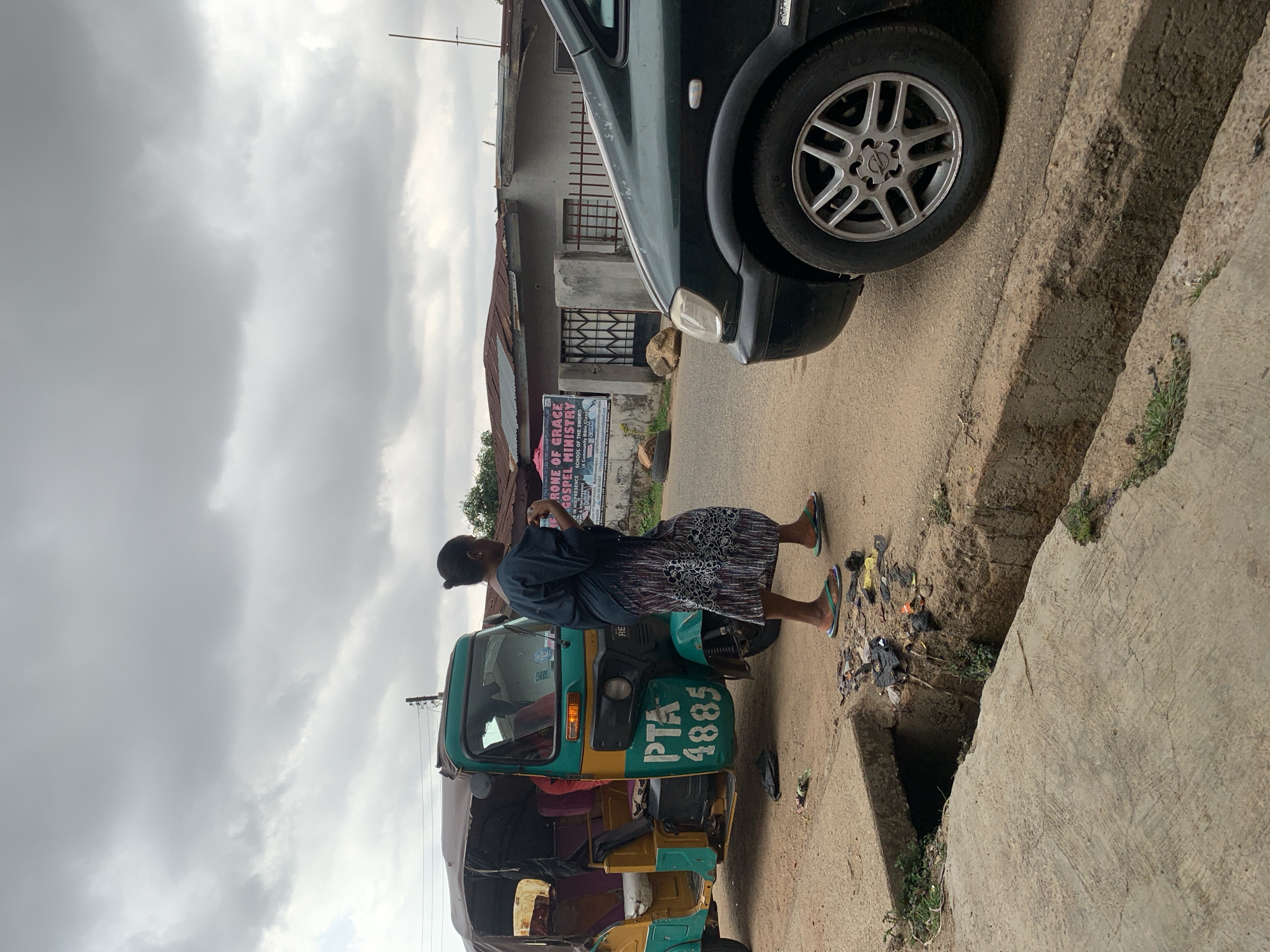 A street scene in Gura Top, Nigeria, under an overcast sky. A woman stands in the street next to a green and yellow motorized tricycle ("keke"), number plate "PTA 4885". She is wearing a dark top and patterned skirt, and sandals. A dark green car is parked near the tricycle. The scene appears to be in front of a building housing the "Rone Of Grace Gospel Ministry". A concrete drainage ditch runs along the side of the road. Litter is visible near the ditch.