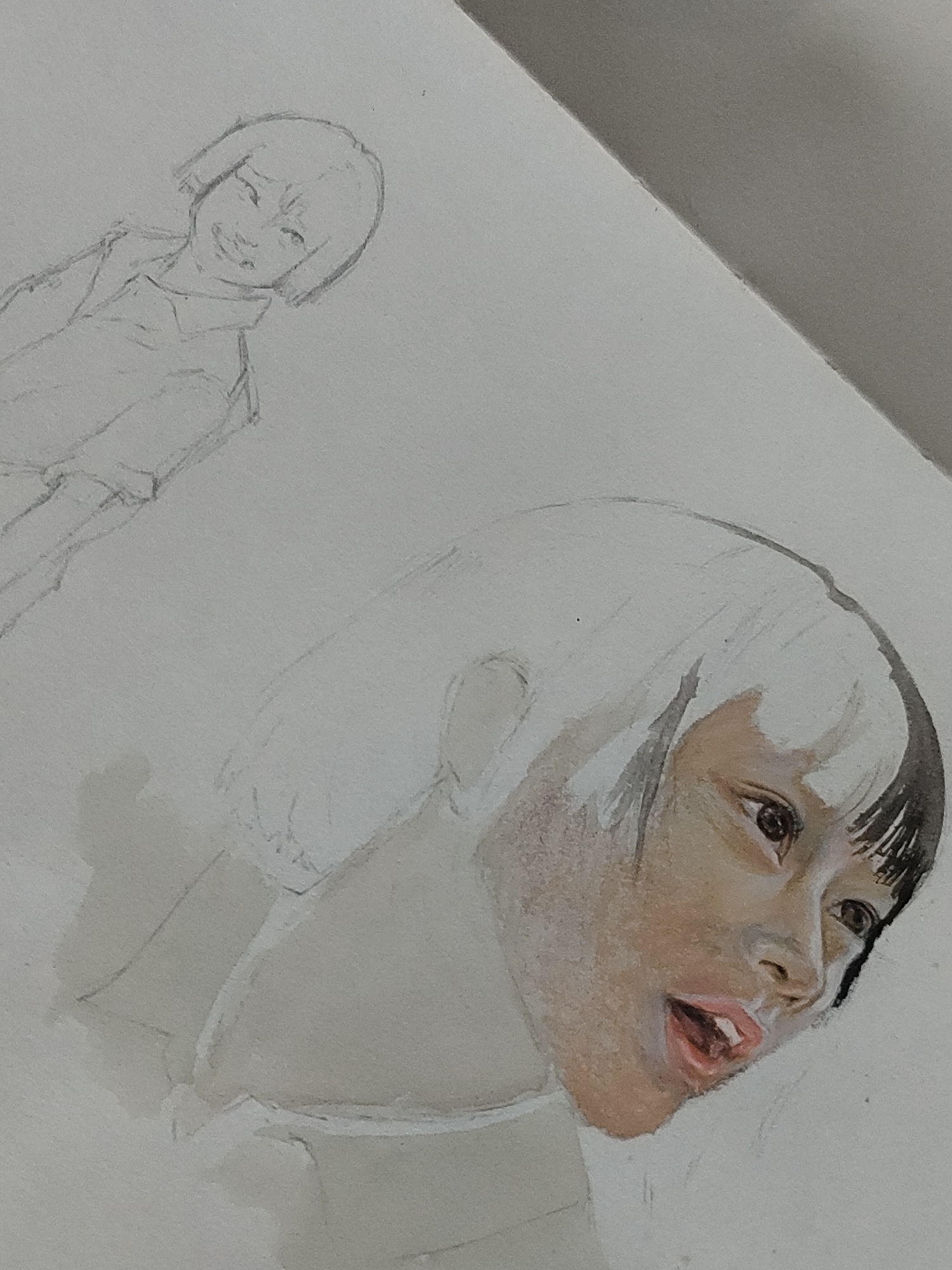 The image displays an angled overhead view of an artistic drawing rendered on a sheet of white, textured paper. The central focus is an expressive, partially colored portrait of a young individual, likely female, located in the lower right portion of the paper. This portrait features a medium-dark skin tone, short black bangs, and a distinct white or light-colored section of hair on the upper head. The subject's mouth is slightly open, revealing teeth and a pink-red tongue, while the wide eyes gaze slightly upwards and to the right. The rendering style suggests watercolor or colored pencil, with notable areas on the left side of the face and upper head remaining uncolored.
Above and to the left of this detailed portrait, a smaller, uncolored pencil sketch of a second individual is present. This sketch depicts a full-body view of a person, also appearing young and likely female, characterized by short hair and a smiling expression. The figure is drawn wearing a collared shirt.
The top right corner of the overall image is occupied by a flat, solid grey surface, which is sharply delineated from the white drawing paper by a diagonal line. The drawing itself does not contain any discernible visual information to establish a specific location within Davao City, Philippines, or any other geographic area.