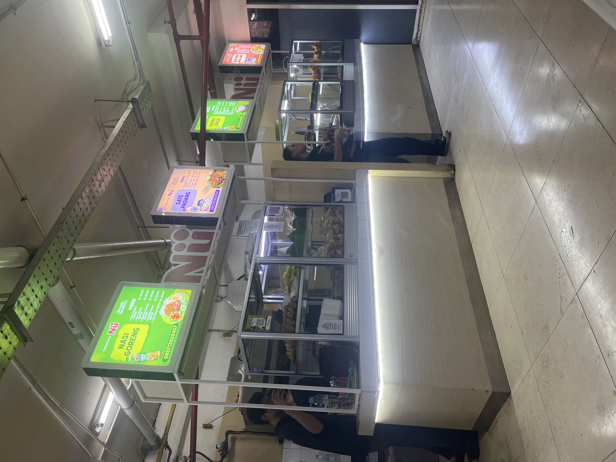 The image depicts a food stall or restaurant interior in South Jakarta, Indonesia. Multiple illuminated signs display menus for various Indonesian dishes, including "Nasi Goreng," "Sate Padang," "Gudeg Jogja," and "Bakwan Malang." The stalls feature glass display cases with food items and stacks of plates and napkins. Two individuals are visible; one, wearing a black shirt and dark pants, appears to be an employee standing behind a counter and interacting with a customer. Another individual, also in dark clothing, is seen from the waist down. The environment suggests an indoor commercial space with tiled flooring, exposed ceiling infrastructure with lighting, and metal shelving units.