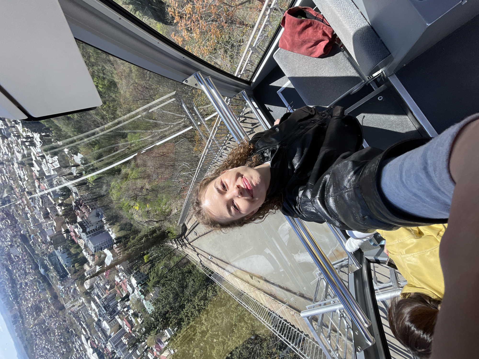 A woman is sitting in a gondola, possibly on a cable car line. Her face is visible and she is looking at the camera with a slight smile. She is wearing a black leather jacket and blue jeans. The gondola has grey seats and metal railings. Outside the window, a cityscape is visible. The scene may be of the city of Tbilisi, Georgia. The season is likely Autumn, because the leaves are changing.