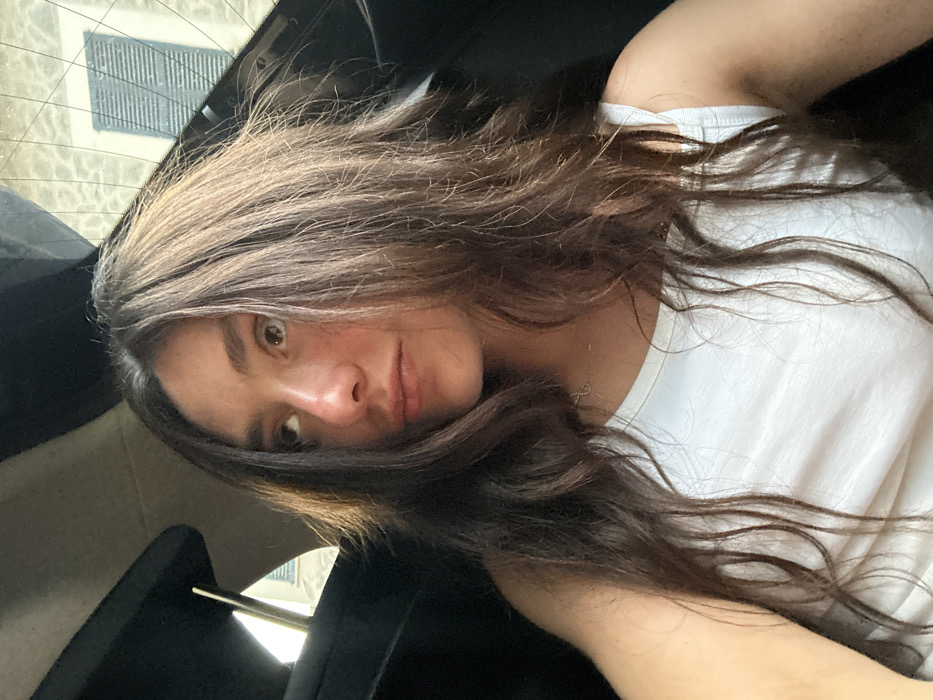 The image features a young individual, likely female, with long, dark brown hair, seated inside a vehicle. The subject is wearing a white sleeveless top and a delicate, gold-colored chain necklace with a small circular pendant. Her gaze is directed upwards and towards the left side of the frame. The interior of the vehicle is dark, with elements such as a headrest and roof lining visible. Through the rear window, a light-colored building exterior is discernible, featuring a window with distinct horizontal slatted blinds. The scene is consistent with a daytime setting in the city of Deià, Spain.
