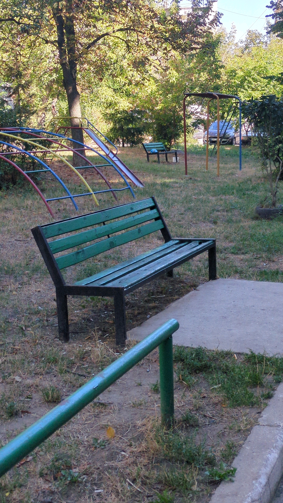 The image captures a playground scene in Chișinău, Moldova. In the foreground, a weathered green wooden bench with a black metal frame sits on a concrete patch. Behind the bench, a colorful, multi-level slide structure with red, yellow, and blue metal bars is partially visible, suggesting children's play. Further back, a swing set with red and yellow supports stands amidst overgrown grass and trees. A dark-colored car is parked in the background, indicating the proximity of a residential area. The overall impression is one of a slightly neglected but functional urban playground, likely part of a residential complex in Chișinău.