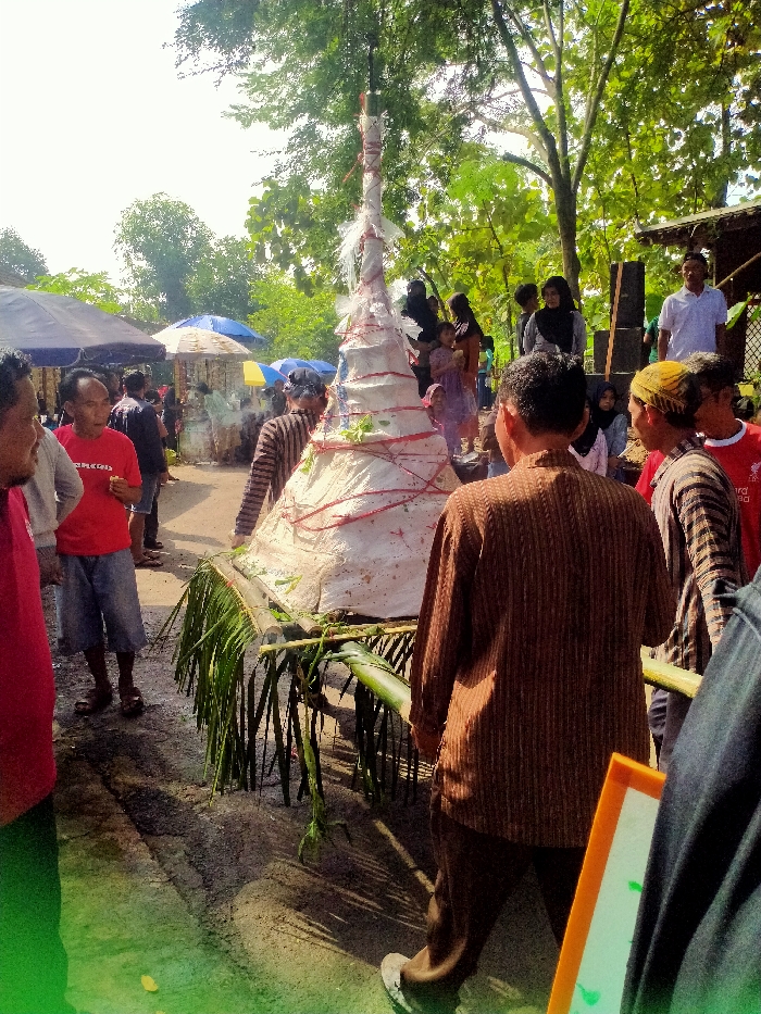 The image shows a group of people gathered around a large white conical structure, located at coordinates -7.471, 110.957 in Masaran, Indonesia. The structure, decorated with red ribbons and green plants, is being carried on a wooden frame by two men. It rests on a bed of palm leaves. The scene is likely a religious or ceremonial event, with several people wearing traditional clothing. The background shows a paved road, green trees, and a simple, partially obscured building. The atmosphere is one of celebration and reverence.