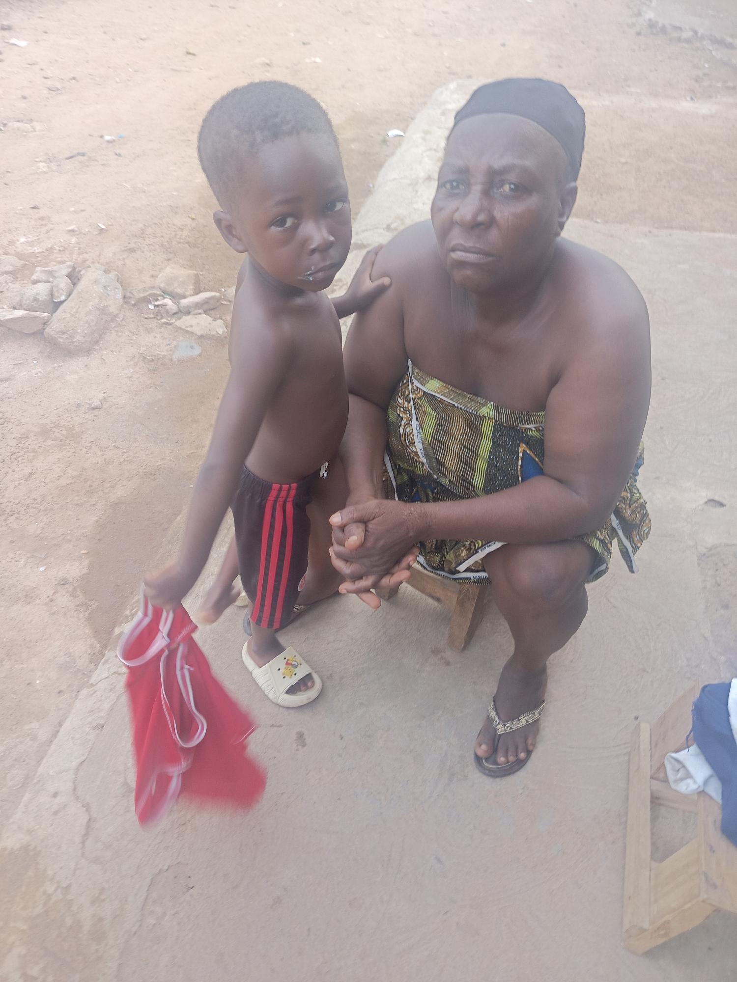 A top-down shot shows a middle-aged Black woman and a young Black child in an outdoor setting. The woman is squatting on a low, simple wooden stool. She is wearing a dark cap or head covering and a patterned cloth wrapped around her chest, leaving her shoulders bare. She has dark skin and a concerned expression. The child stands to her left, partially obscuring her. The child is wearing black shorts with red stripes, one sandal, and holding a red shirt in their hand. The child has a smudge of white on their face near their mouth. The background consists of a rough, textured surface, possibly concrete or packed earth. In the distance, there is a dirt area with rocks and sparse vegetation. The location is Akwanga, Nigeria.