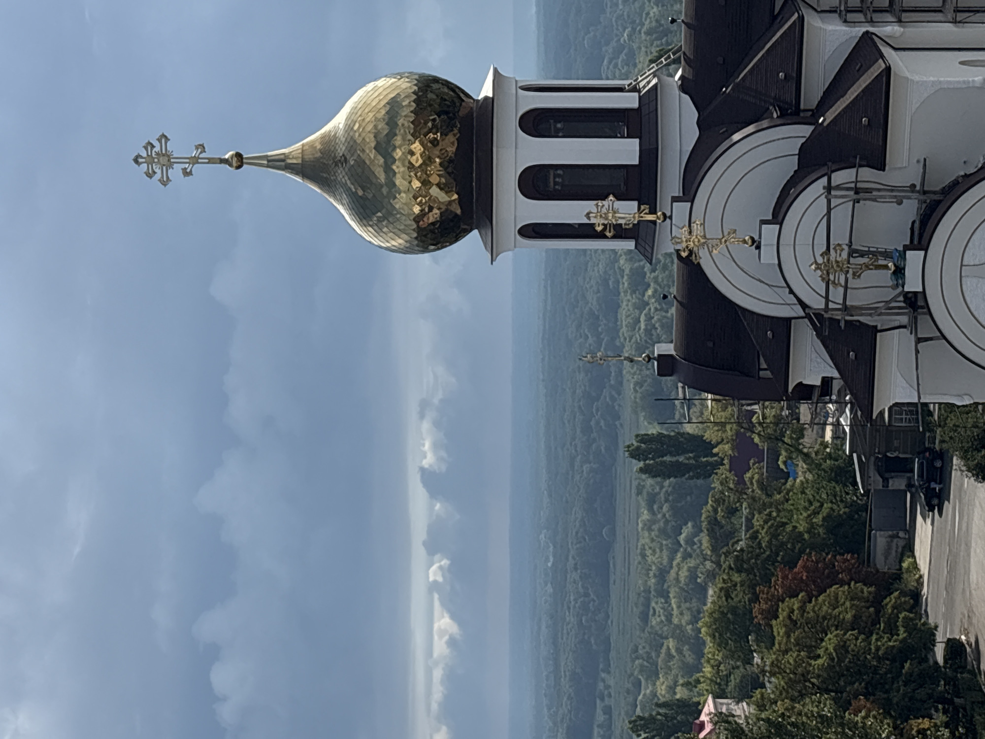 This image shows a close-up view of a golden dome of an Orthodox church, possibly located in Bobrov, Russia. The dome is intricately patterned and topped with a multi-tiered cross. The church architecture features white walls, dark roofs, and several smaller crosses. In the background, a lush green forest stretches to the horizon under a dramatic sky with heavy clouds. The overall scene suggests a serene and spiritual setting, possibly captured during or after a period of rain, given the cloudy sky.