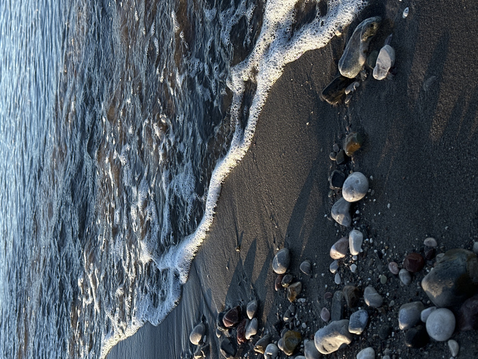 The image depicts a natural shoreline located in Šušanj, Montenegro. The upper right portion of the frame shows a body of dark, rippled water, exhibiting deep blue-grey tones. A distinct, curvilinear band of white, frothy foam extends across the mid-ground, marking the point where water interfaces with the land. The lower left and central sections feature a dark grey, moist sandy beach, densely covered with various smooth, rounded pebbles and rocks. These geological elements range in size and display a spectrum of colors including light grey, white, reddish-brown, dark brown, and black. Elongated shadows are cast by several pebbles onto the sand, indicating a low-angle light source. No human figures, animals, or man-made structures are present within the scene. The primary observed action is the dynamic interaction of water and foam with the static sandy and rocky beach substrate.
