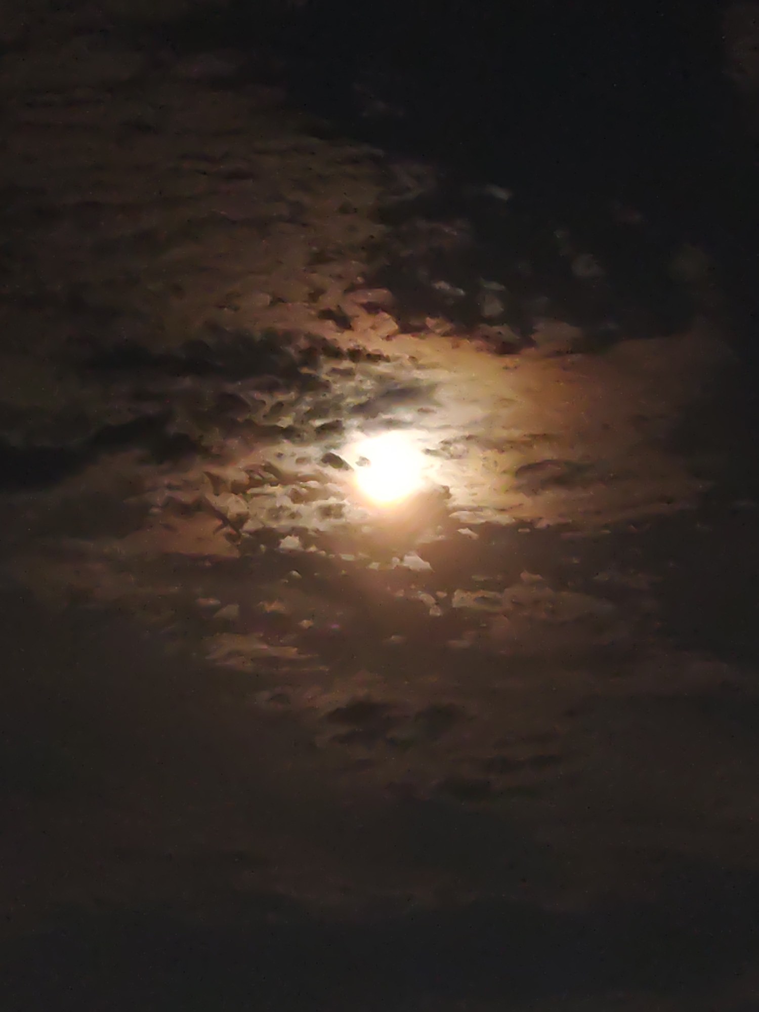 The image is a nighttime photograph of the sky, with the moon partially obscured by clouds. The moon is a bright, glowing orb, emitting a warm, golden light that illuminates the surrounding clouds. The clouds themselves are a mixture of dark, shadowy masses and lighter, wispy tendrils, creating a dramatic and atmospheric scene. The overall impression is one of mystery and ethereal beauty. The photograph appears to have been taken in Petropavl, Kazakhstan, though no specific landmarks or details are visible to confirm this location.