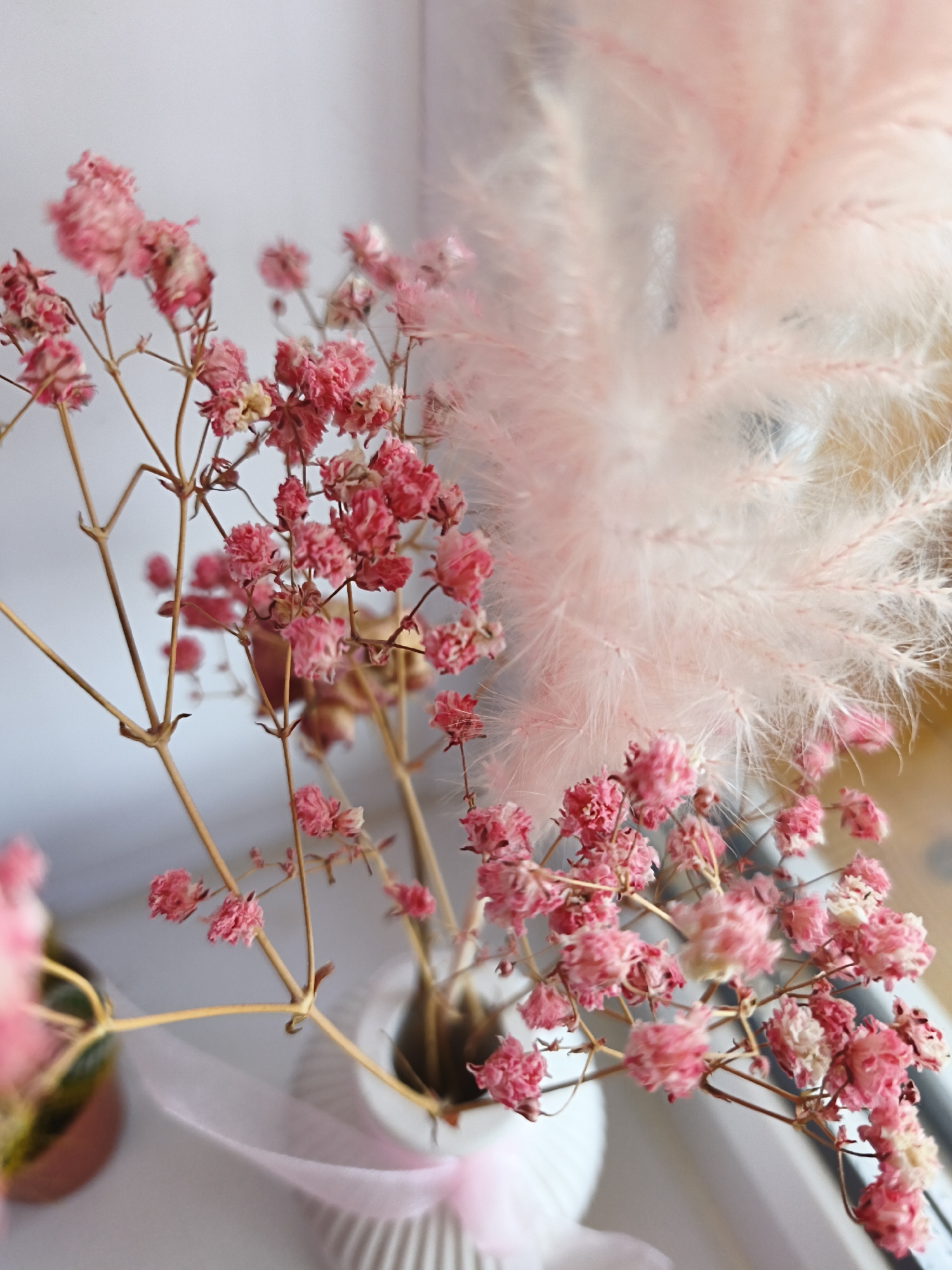 The image shows a close-up of dried pink gypsophila flowers in a white ribbed vase, with a blurred pink feathery plant in the background. A pale pink ribbon is draped across the vase and the surface it sits on. The scene is softly lit, likely by natural light. There are no people or animals visible. The setting appears to be indoors, possibly on a windowsill, given the light and the slight hint of a window frame on the right. The exact location in the city of Naberezhnye Chelny, Russia, is not discernible from the visual information provided in the image.