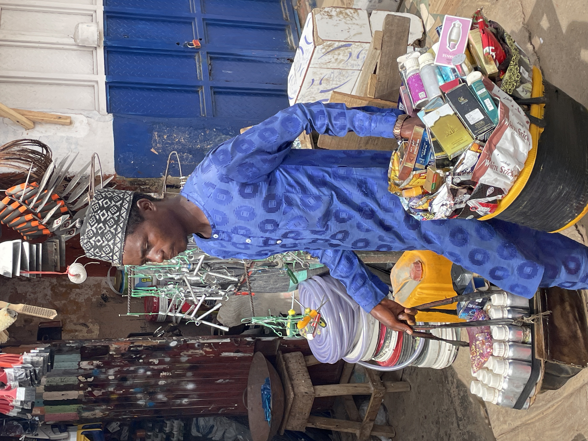 In a street scene, a man wearing a blue patterned shirt and a black and white patterned skullcap stands among a variety of merchandise. He appears to be sorting or examining items, perhaps engaging in trade. The merchandise includes stacks of metal sheeting, coils of plastic tubing, bottles, and a large assortment of packaged goods such as incense sticks and fragrance oils arranged in a cylindrical container. The environment suggests a bustling marketplace, indicative of street commerce in a location that may be Jos, Nigeria. The setting consists of painted metal doors, stacks of tiles, and various tools and hardware, indicating a commercial or industrial district.