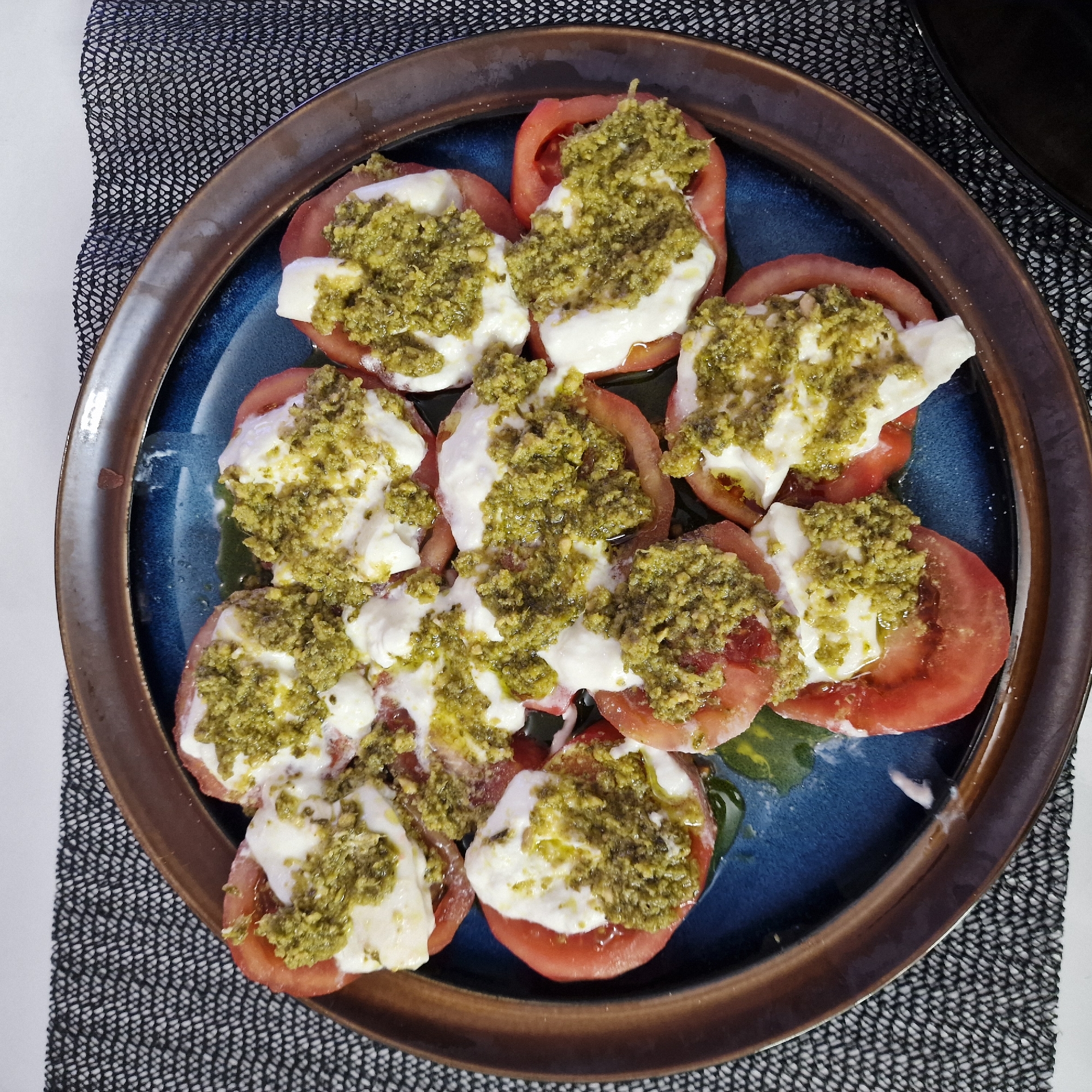 A top-down shot shows a plate of food, likely Caprese salad, sitting on a textured mat. The plate is blue with a brown rim and contains sliced tomatoes topped with white cheese (possibly mozzarella) and green pesto. The mat is patterned in black and white, placed partially on a white surface. There are no people visible in the image. No information about the location in Sofia, Bulgaria is discernible from this image.