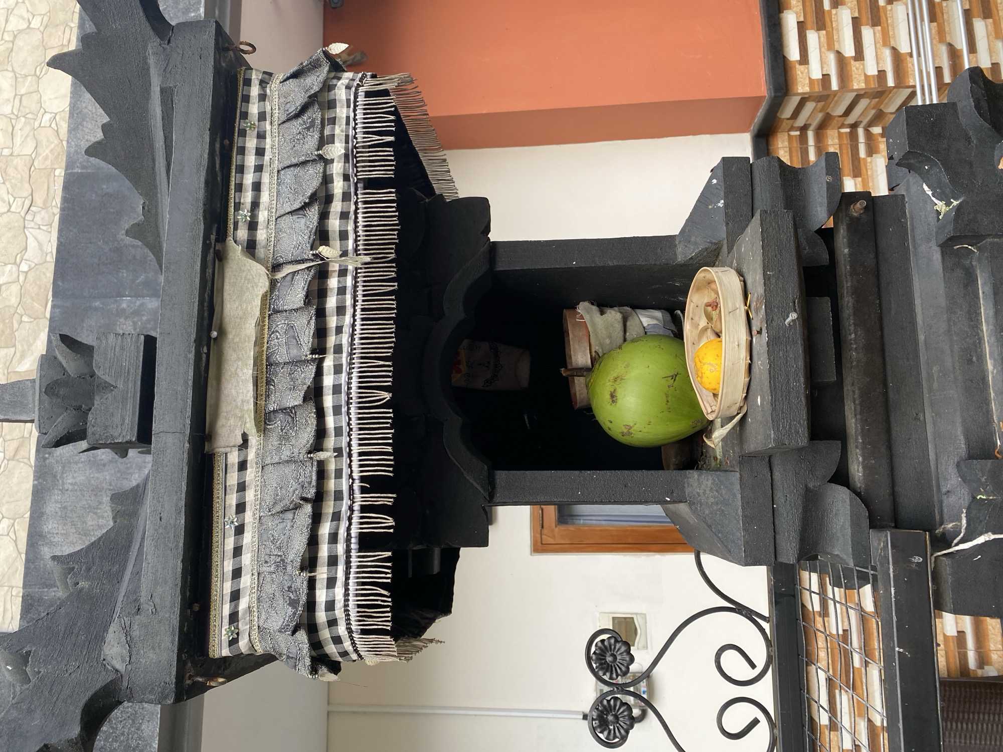 The image captures a cultural artifact in Nusa Dua, Indonesia, specifically a Balinese offering shrine, commonly found in local households or businesses. The shrine is constructed from carved black wood and is adorned with black and white checkered fabric featuring fringe detailing. Inside the shrine, offerings are visible, including a green coconut and a yellow fruit or flower in a woven basket. The shrine's background includes a white wall, an orange roof, and a tiled surface. The presence of an ornate wrought iron fence is also evident.
