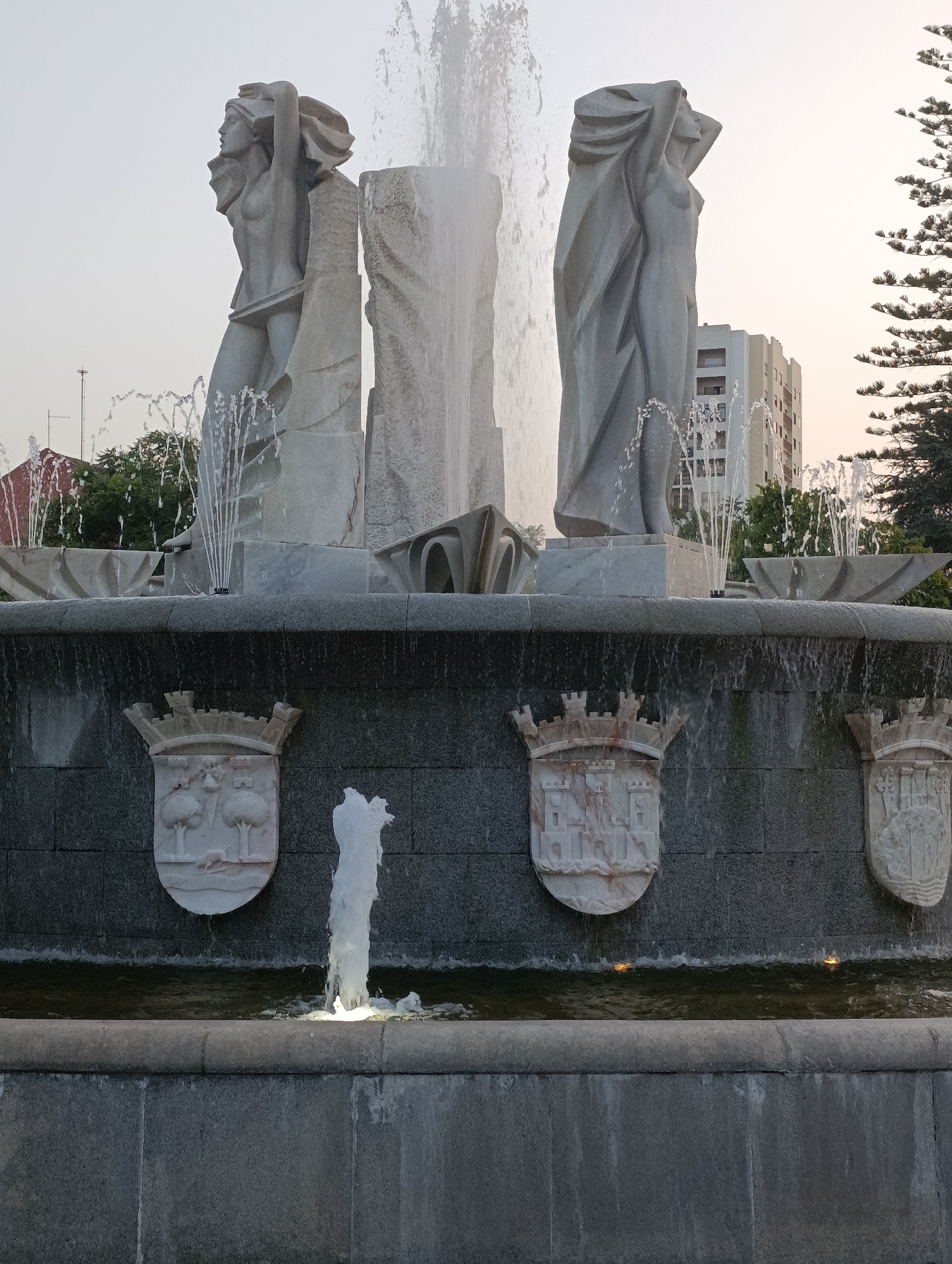 The image depicts the Avenida Luísa Todi Fountain in Setúbal, Portugal. The fountain features two large, stylized human figures sculpted from marble, flanking a central water feature. Smaller jets of water spray upwards around the base of the sculptures. The main basin of the fountain has a dark, textured wall adorned with three marble escutcheons, each bearing a coat of arms. Water cascades over the rim of the basin, creating a shimmering effect. In the background, a tall, modern building and trees are visible against a pale sky, suggesting an urban setting. The lighting indicates it is either early morning or late afternoon.