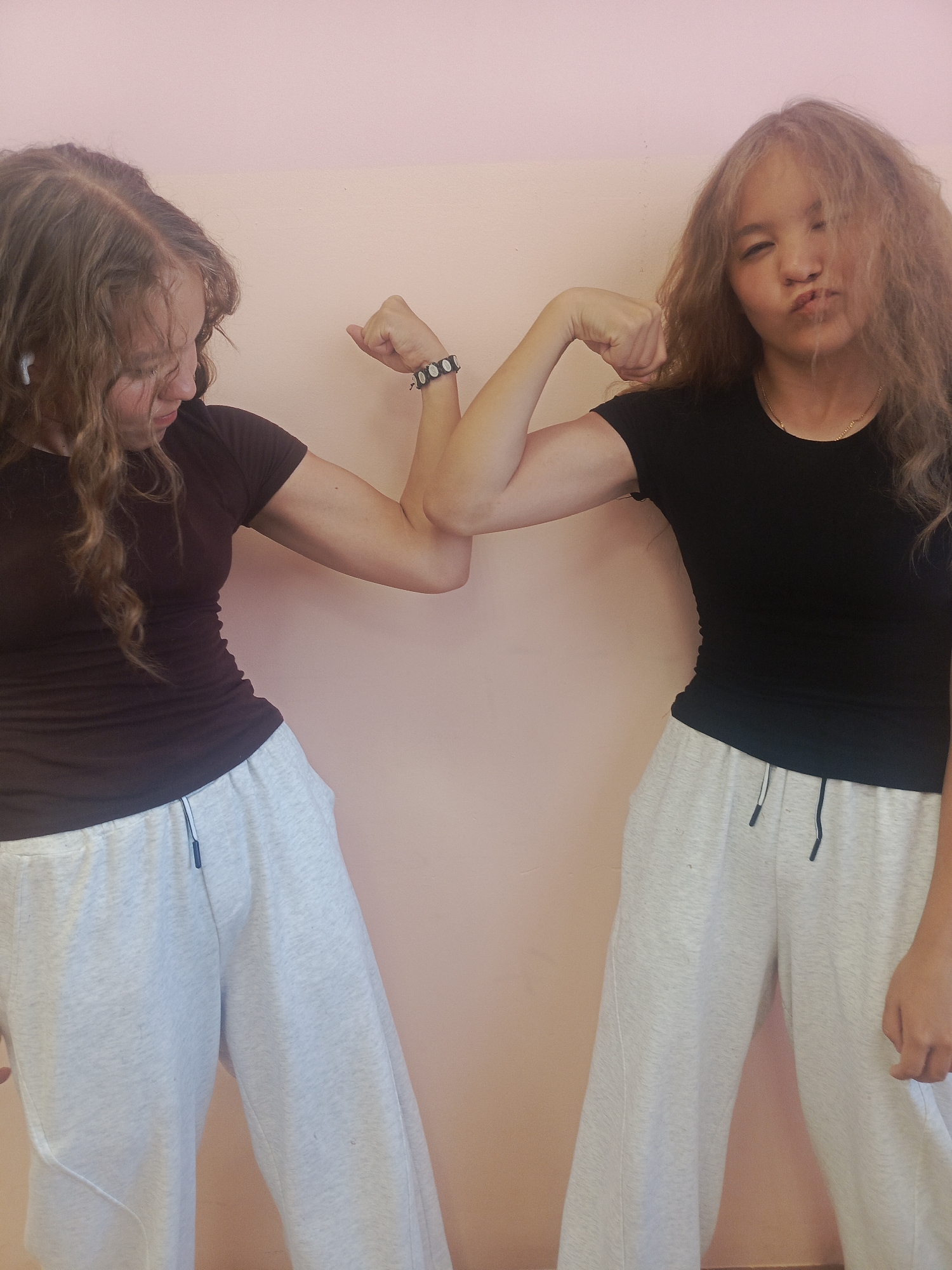 Two young women stand side-by-side against a pale pink wall. The woman on the left has brown, curly hair and is wearing a dark maroon t-shirt and light gray sweatpants. She has an earbud in her left ear and is flexing her left arm, looking down at her bicep. She is wearing a bracelet. The woman on the right has wavy, light brown hair with blonde highlights and is wearing a black t-shirt and light gray sweatpants. She is also flexing her left arm and is making a pouting face. The scene appears to be indoors.