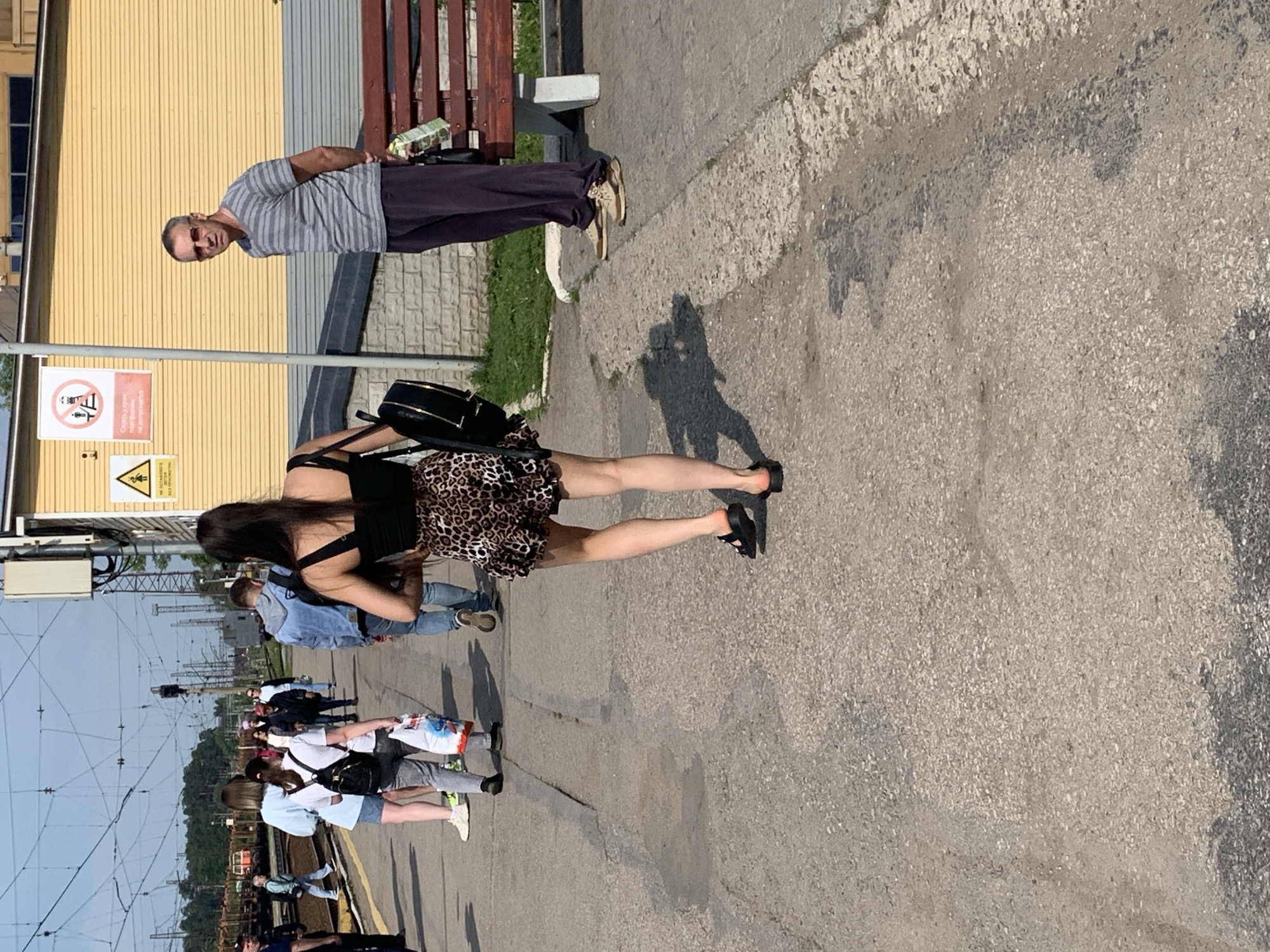 In a sunlit outdoor scene, likely a train station platform in Kungur, Russia, a group of people are gathered. The ground is paved with asphalt and gravel. In the foreground, a woman with dark hair, wearing a black crop top and a leopard-print skirt, walks away from the camera, carrying a black backpack. To the right, a man with short gray hair, wearing sunglasses, a striped t-shirt, and dark trousers, stands looking towards the left, holding a carton of juice. Behind them, several other individuals are visible walking along the platform, some carrying bags. A yellow building with a corrugated metal lower section runs along the right side of the platform, and a wooden bench with metal legs is situated in front of it. Overhead, a complex network of electrical wires crisscrosses the sky. A yellow warning sign with a black border depicting a person and a triangle is mounted on a pole near the building.