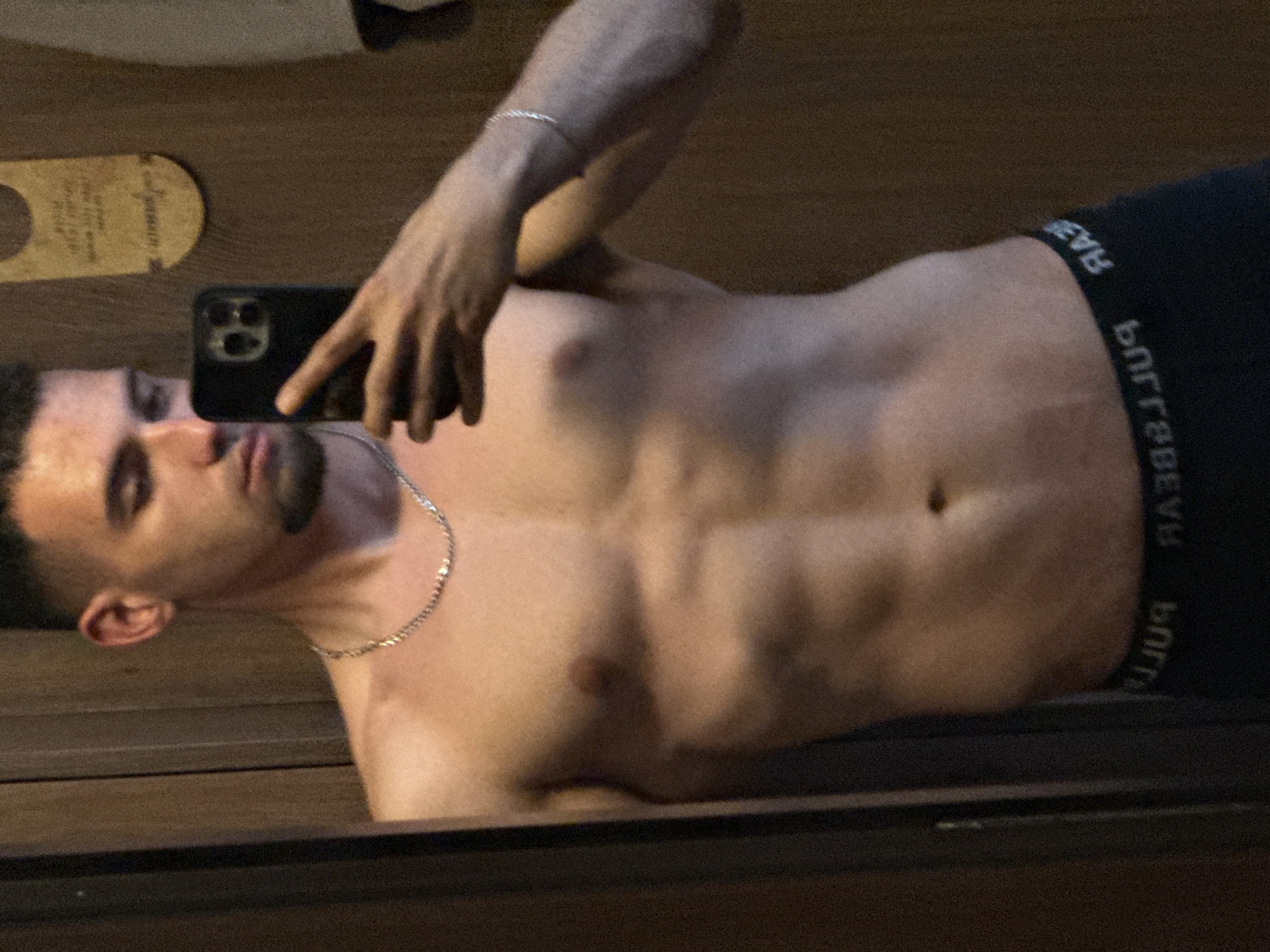A shirtless man is taking a selfie with a black smartphone. He has a goatee, a silver chain necklace, and a silver bracelet. He is wearing black underwear with white lettering on the waistband. He is standing in front of a mirrored surface that reflects a wooden surface. A hotel room key card is on the left side of the frame. It is not possible to determine the location in the city of Veliko Tarnovo, Bulgaria, from the provided image.