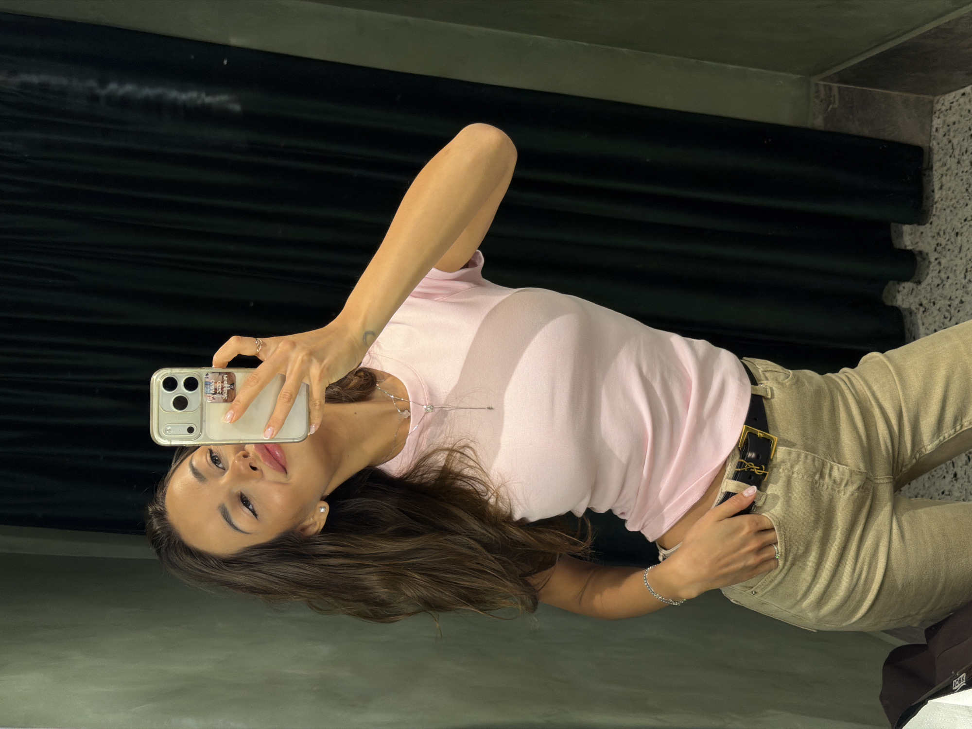 A young woman with long, brown, wavy hair takes a selfie in what appears to be a fitting room. She is wearing a light pink t-shirt, beige pants, and a black belt with a gold buckle. A silver necklace and bracelet adorn her neck and wrist. Her left hand is in her pocket, while her right hand holds up her phone to capture the reflection. The background consists of a muted green wall and dark green velvet curtains. The floor is speckled with gray and white. There is no indication that this image was taken in Saint Petersburg, Russia.