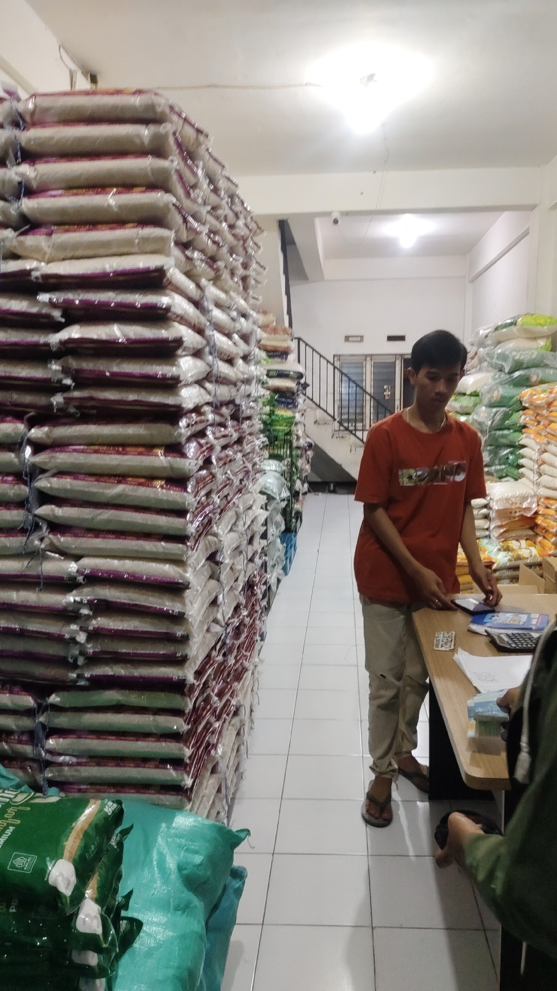 In Malang, Indonesia, a young man in an orange t-shirt and khaki pants stands behind a wooden counter in a store filled with sacks of rice. The sacks are stacked high on shelves on both sides of the narrow aisle. The man is looking down at a tablet device on the counter, and he is holding a small stack of money in his left hand. A calculator and some papers are also on the counter. Another person, whose face is not visible, is partially seen on the right side of the frame, holding a hand out towards the counter. The floor is tiled with white squares. The scene suggests a retail or wholesale environment for rice.