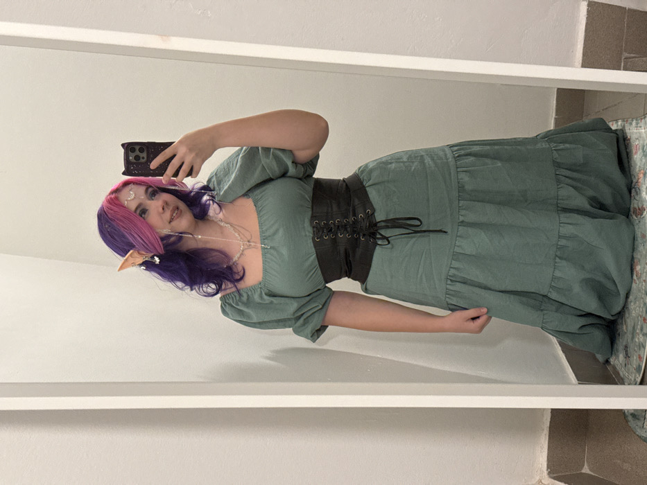 A full-body mirror selfie captures a young person dressed in a fantasy-inspired outfit, likely taken indoors against a plain light-colored wall. The person, who appears to be female, is smiling gently and looking towards the camera (their reflection in the mirror).
Her hair is a striking blend of vibrant colors, with a bright pink at the roots and front transitioning to a deep purple along the length. It is long and wavy. Adorning her forehead is a delicate silver-colored headpiece featuring a crescent moon and star motifs, and glitter is visible around her eyes and forehead. She is wearing light-colored, possibly blue, eye makeup, and light-toned elf ears are visible peeking out from under her hair. A long, layered necklace made of small, clear beads drapes over her chest.
She is wearing a long, light greenish-blue (resembling sage green) dress. The dress features short, puffed sleeves and a square neckline. A prominent dark brown or black wide corset-style belt is cinched around her waist, with lacing visible at the front. The skirt of the dress is tiered, suggesting a flowing or voluminous design.
The person is holding a dark-colored smartphone in their left hand, angled to capture the mirror reflection. The phone case has a visible texture or design. Her right hand is relaxed at her side, near the skirt of the dress.
The background reflected in the mirror is simple, consisting of a plain light-colored wall. The lower right corner of the image shows a partial view of a tiled floor (in shades of brown or beige) and a small, patterned rug. The overall impression is that of a prepared look, possibly for cosplay, a themed event, or as a personal style expression. The lighting is even and typical of an indoor setting.